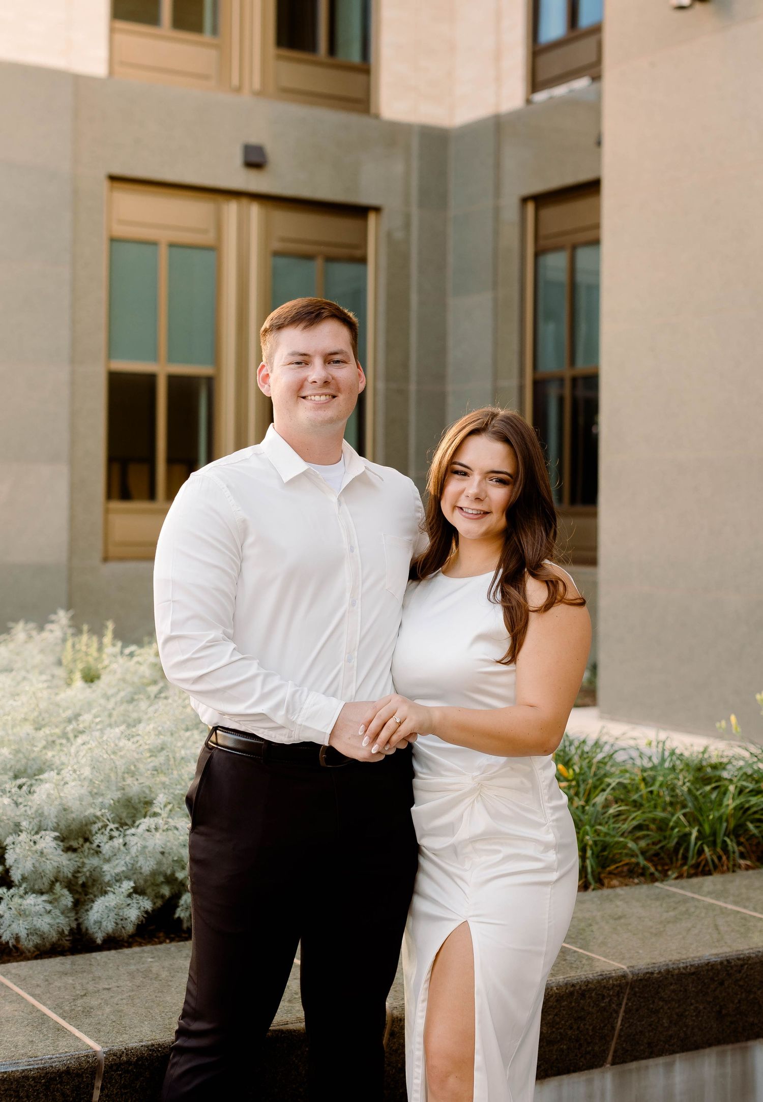Elegant couple in white and black attire poses together outside a modern building.