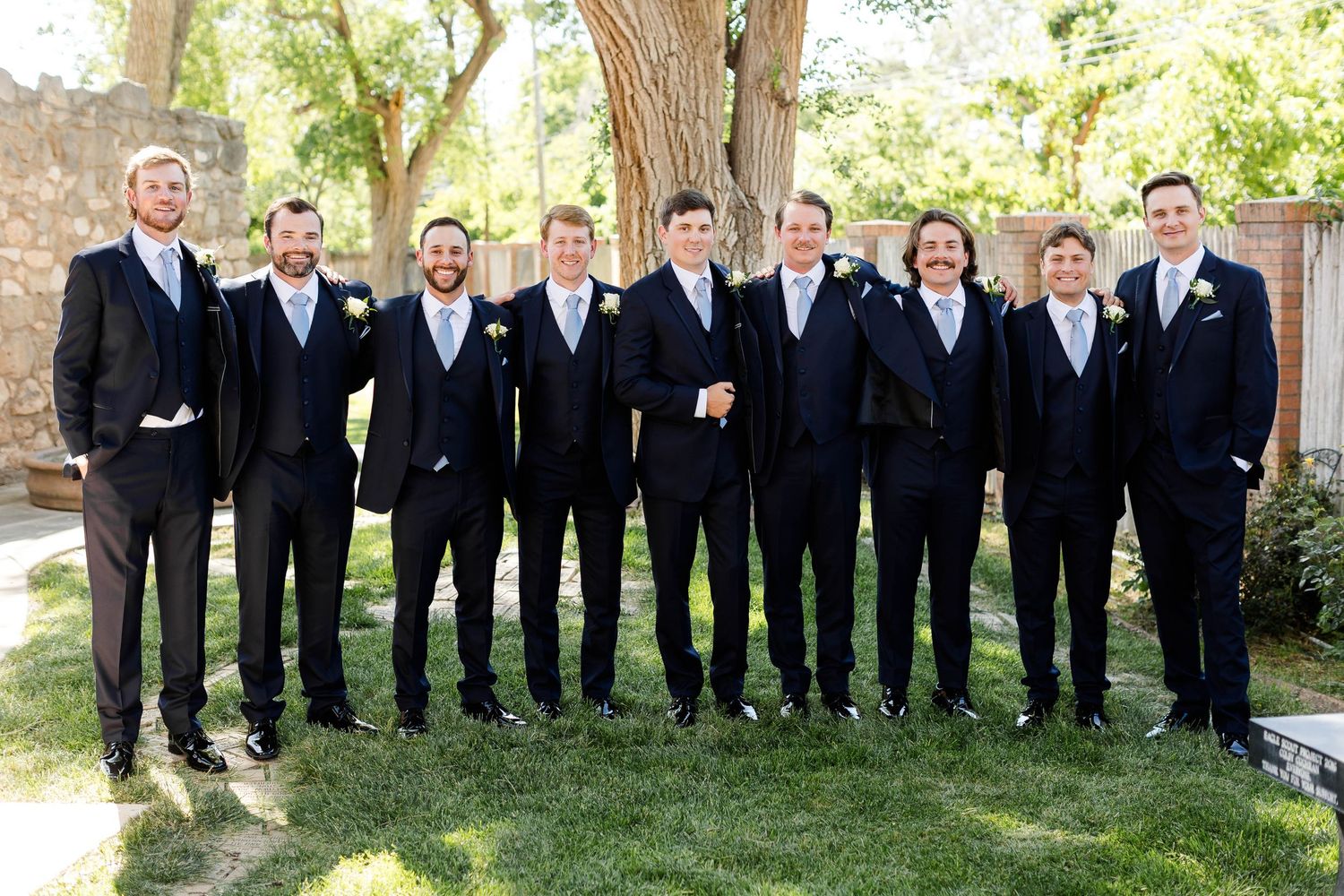 Large group of groomsmen in dark suits lined up outdoors under trees.
