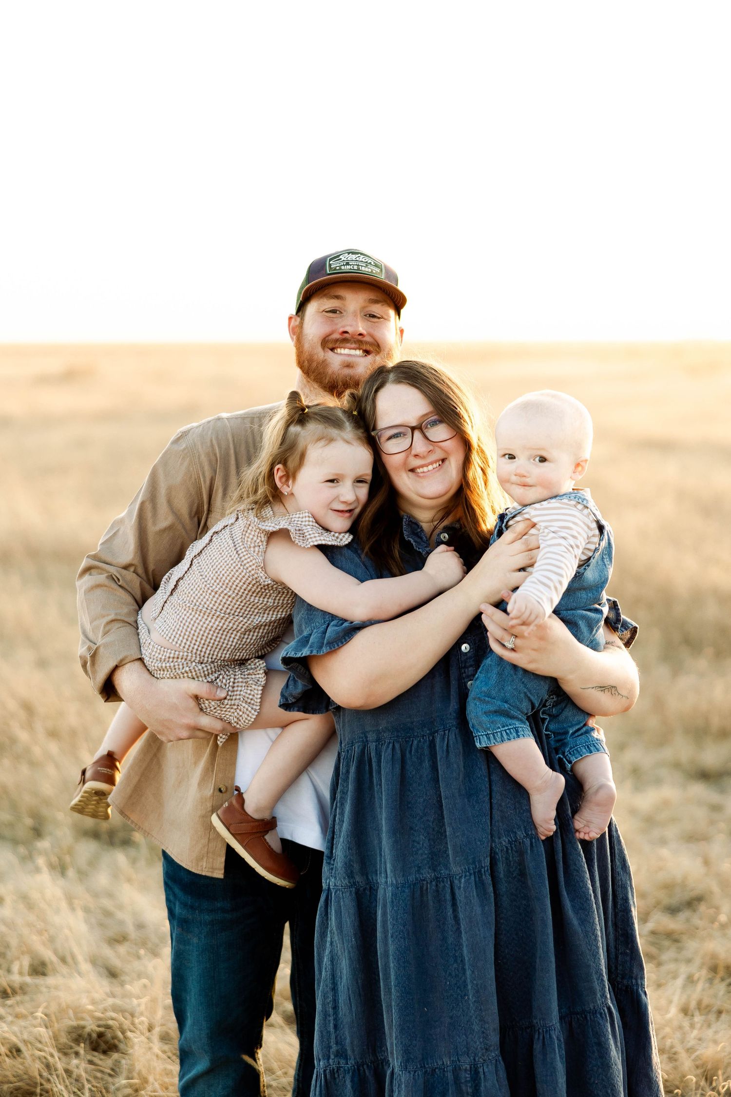 Family of four poses together in a golden field at sunset wearing casual clothing.