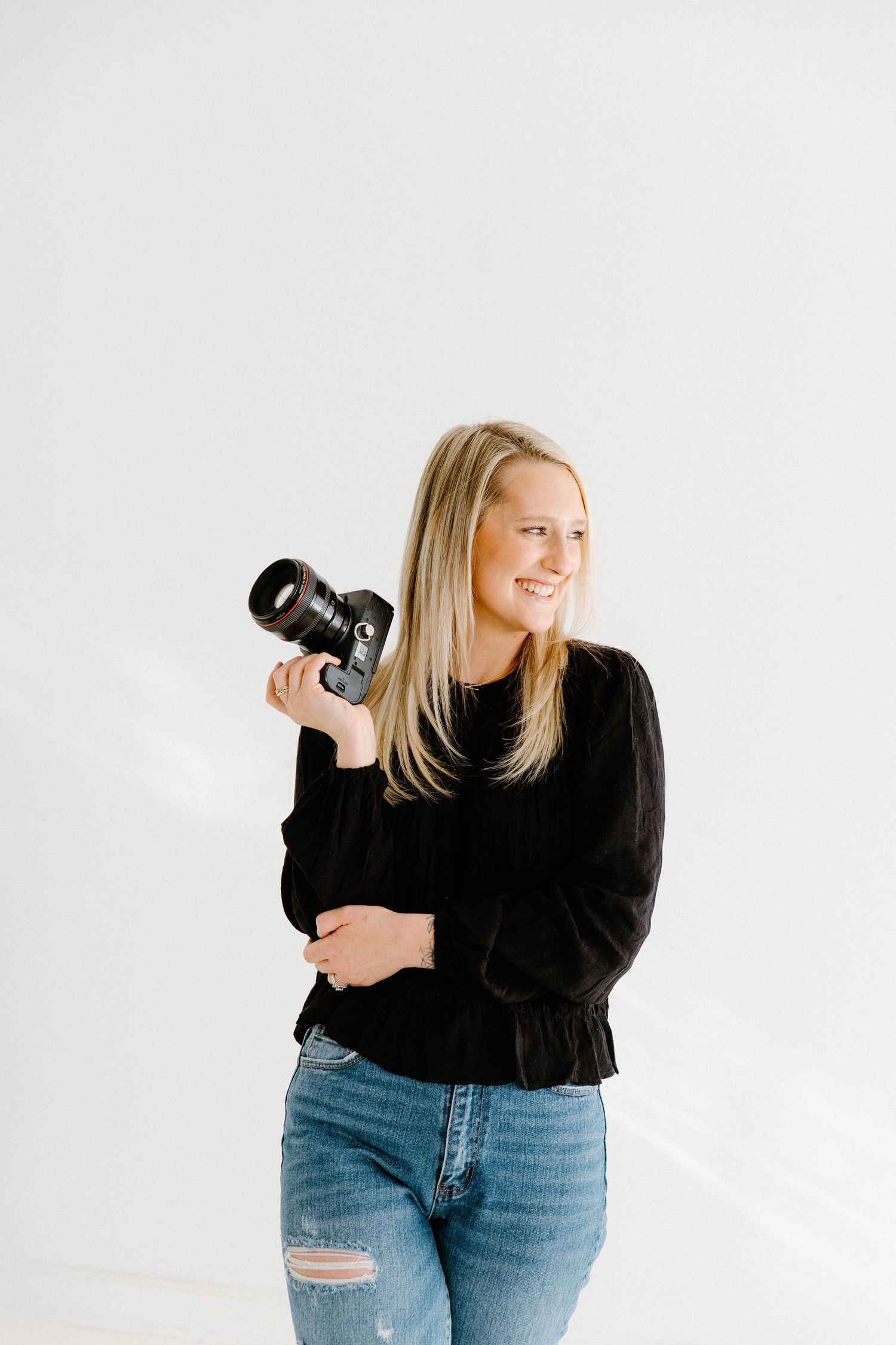 Smiling blonde woman holding a DSLR camera, wearing a black top and jeans, posing in a bright white studio.