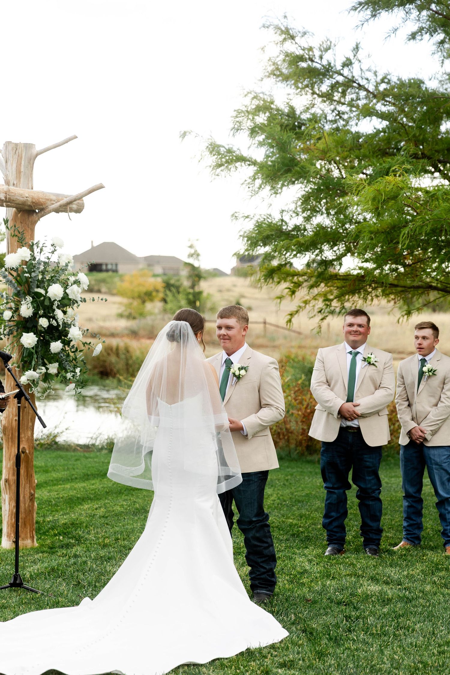 Wedding party poses outdoors on green lawn with rustic wooden arch and white flowers.