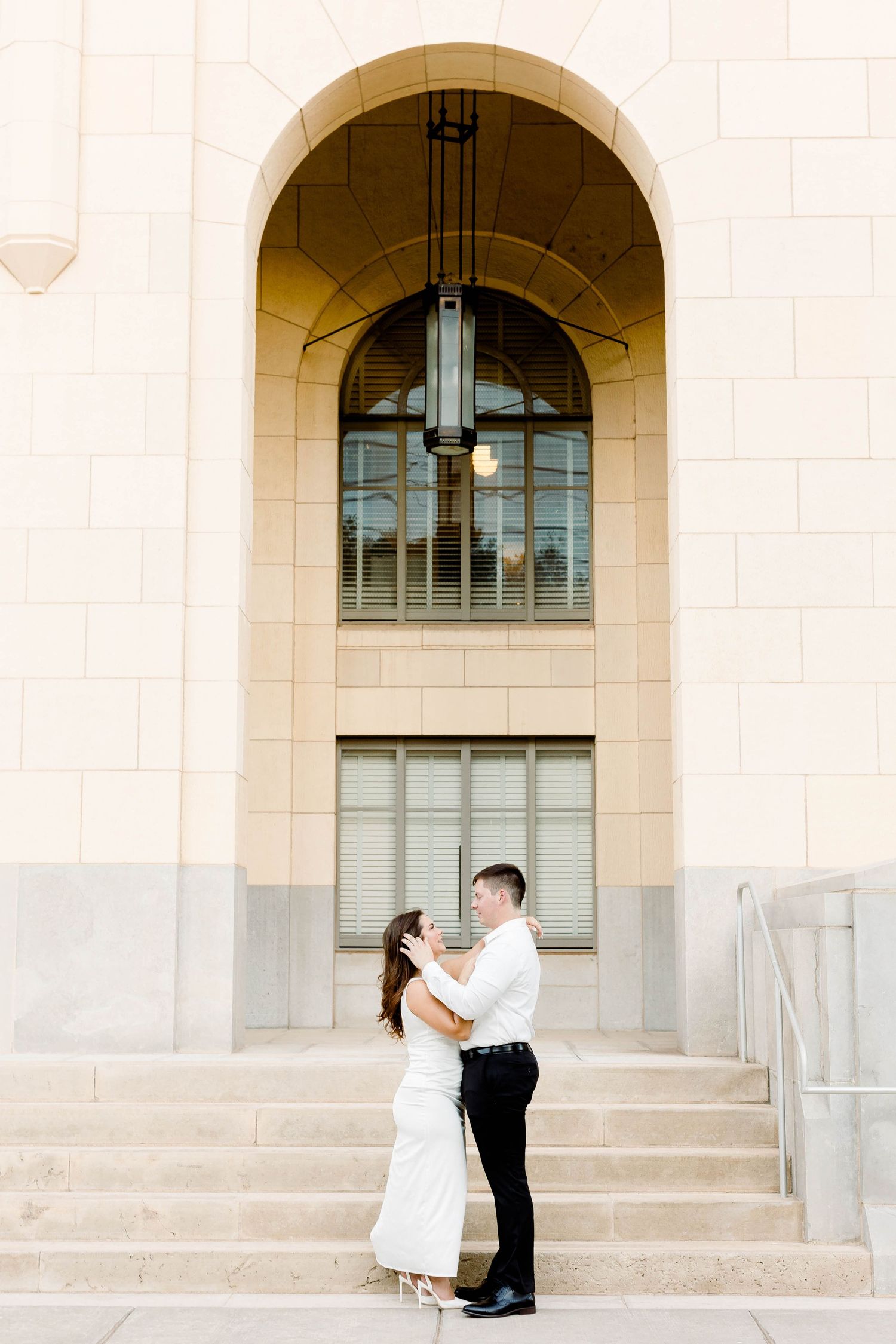 Romantic couple poses on elegant stone steps with arched windows in the background.