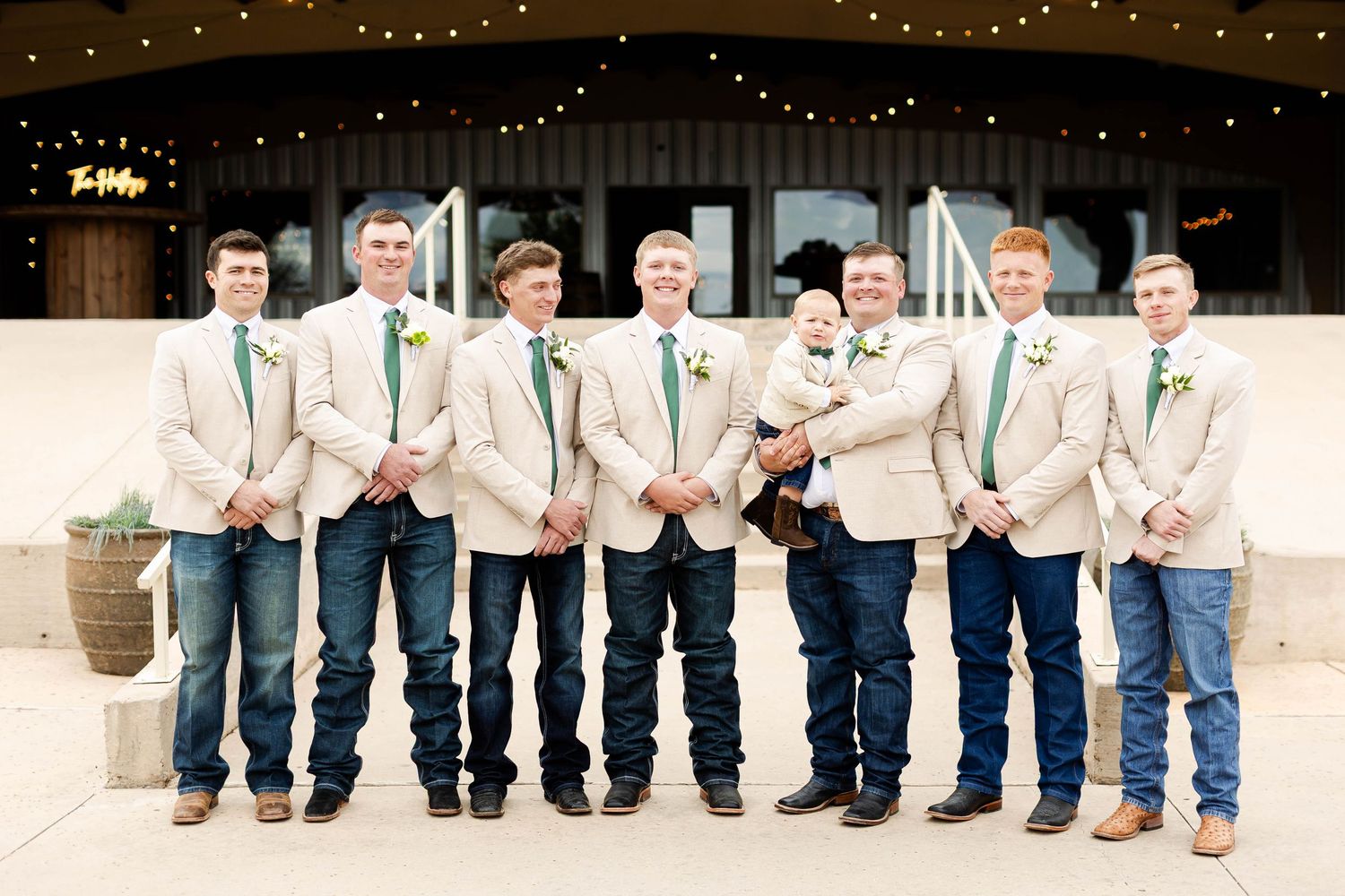A wedding party in jeans and dress shirts stands in a row showing off their brown boots against a rustic backdrop.