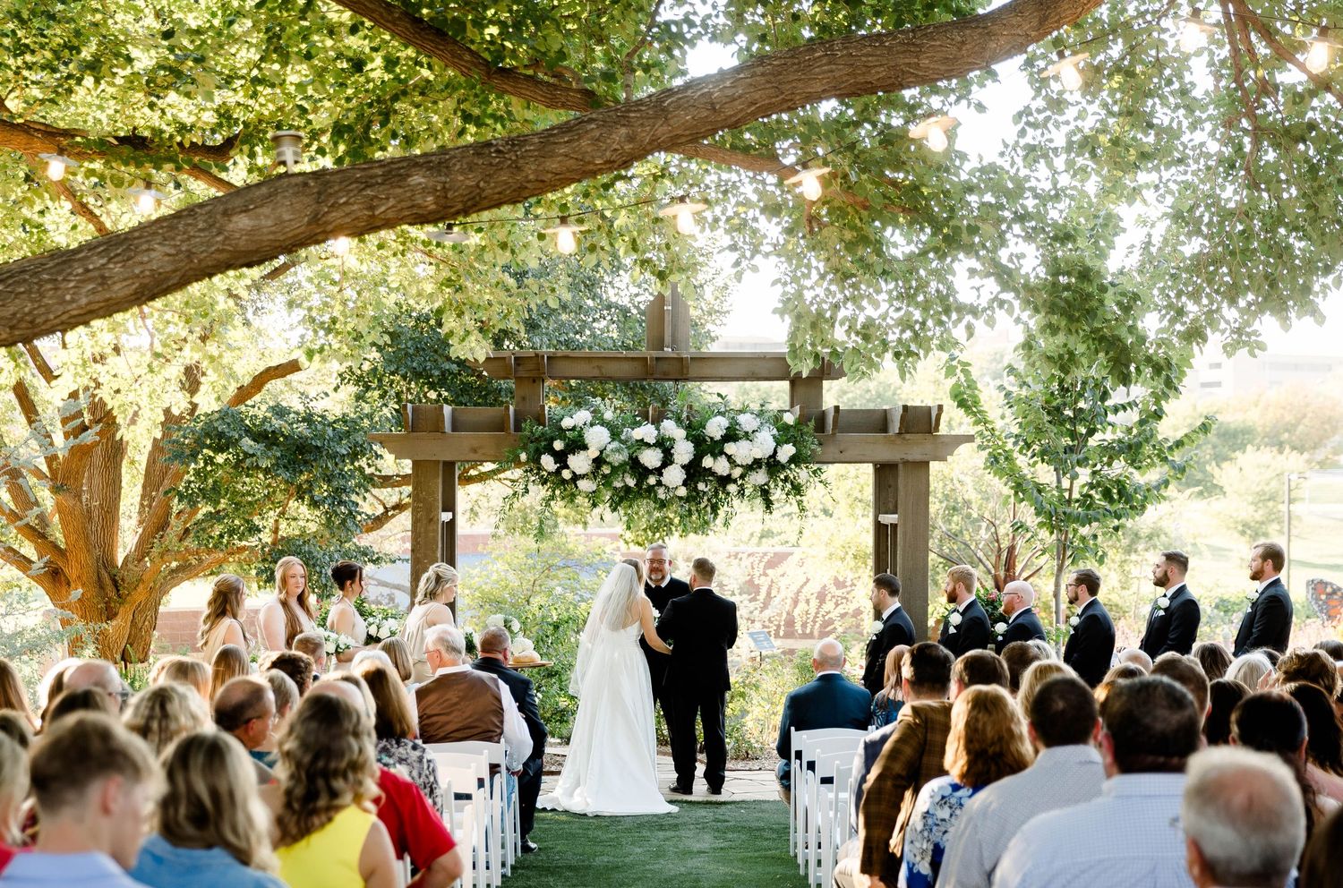 An outdoor wedding ceremony under large trees with white chairs set up for guests.