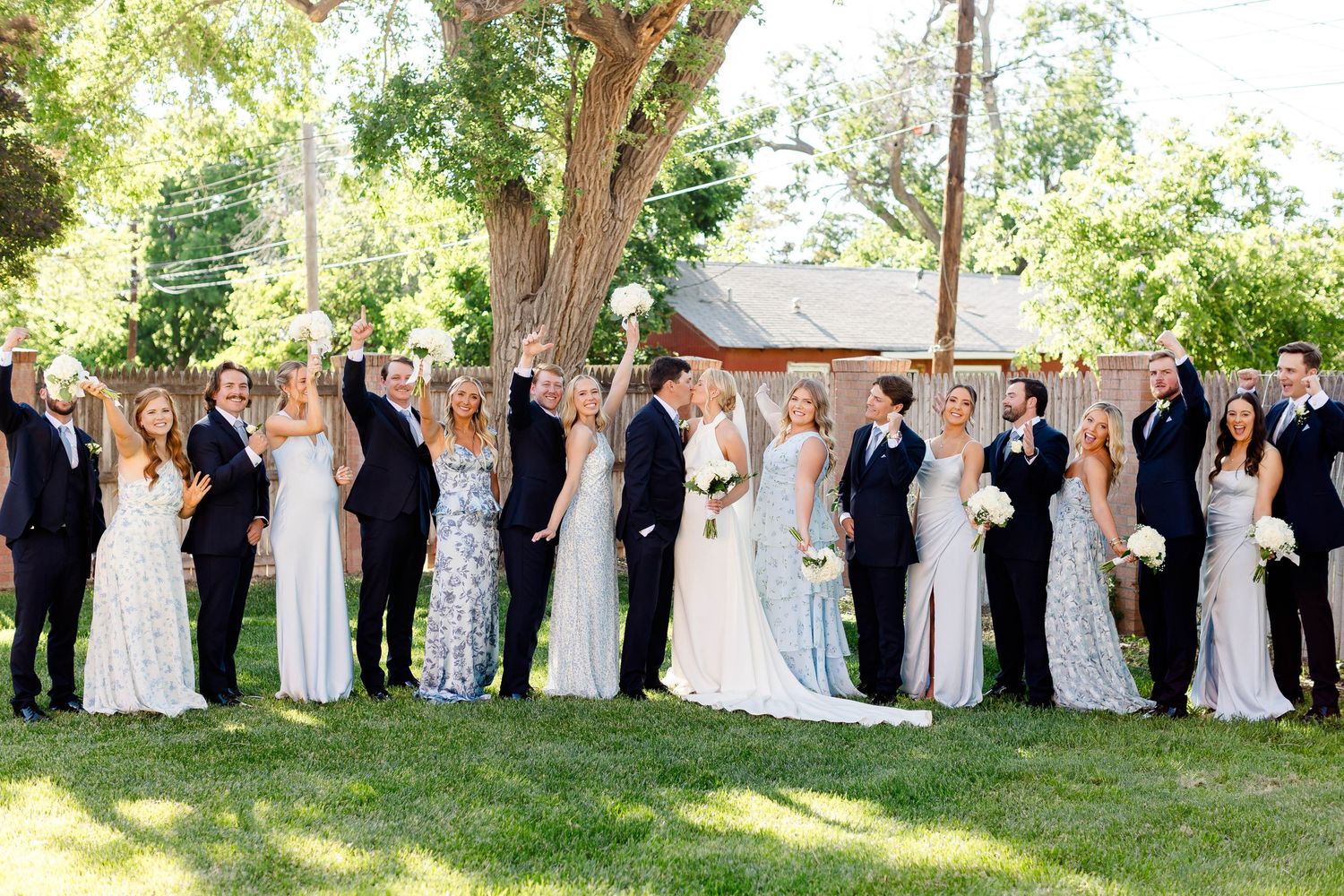 Wedding party members arranged in a line outdoors with bridesmaids in light dresses and groomsmen in dark suits.