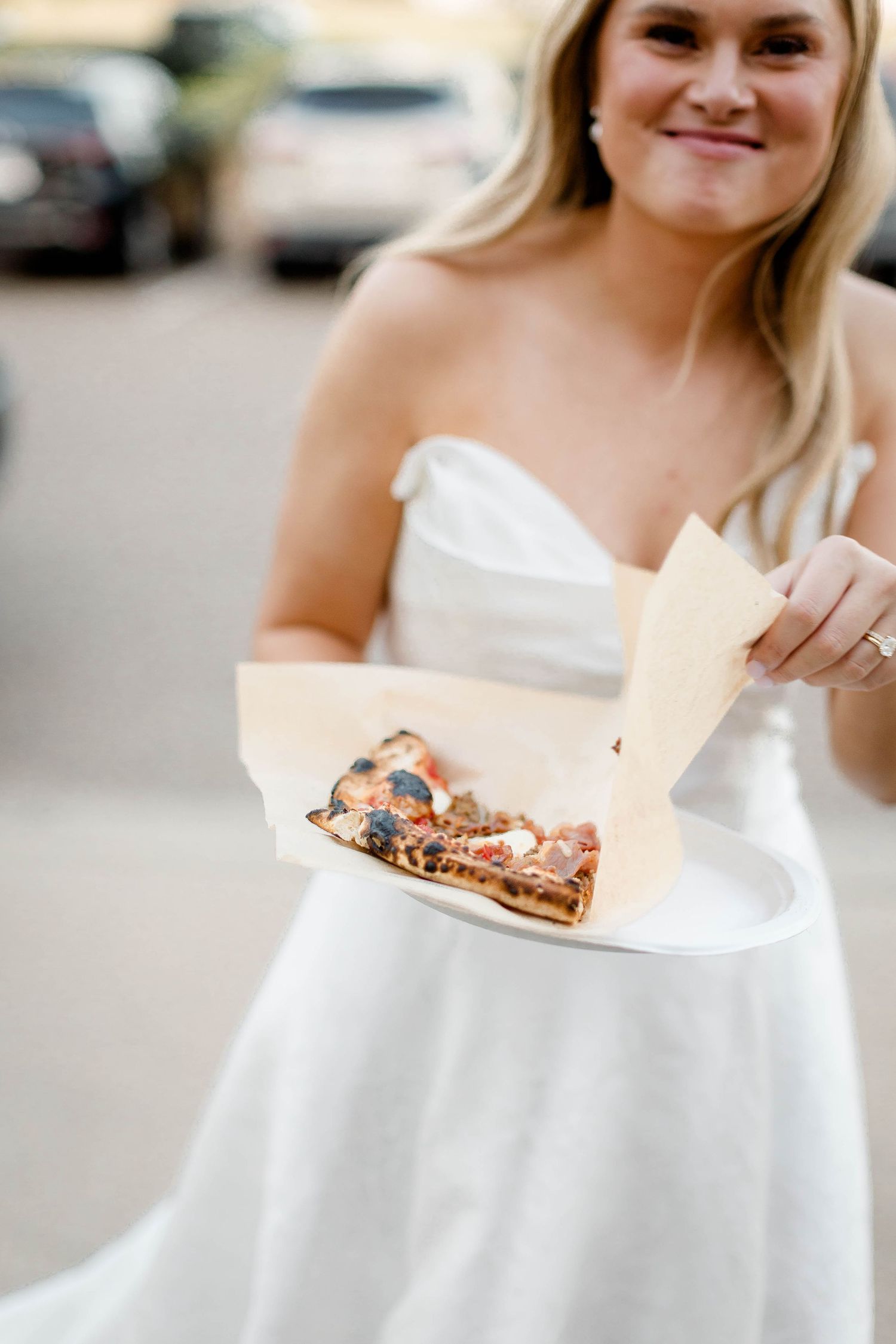 A person in a white dress smiles while holding a takeout container of food at an outdoor event.