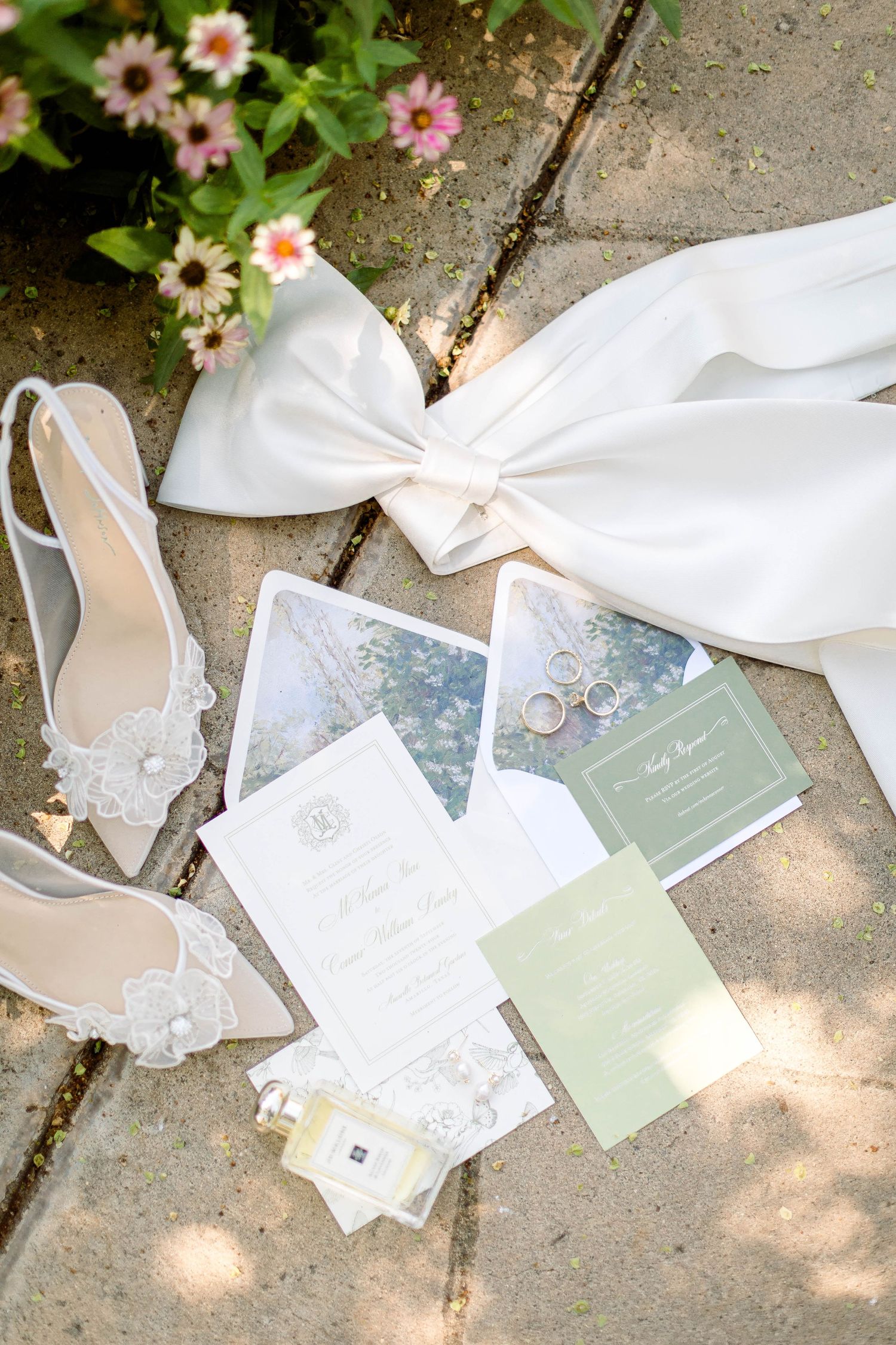 Wedding stationery suite with white and sage green invitations laid on stone ground with pink flowers.
