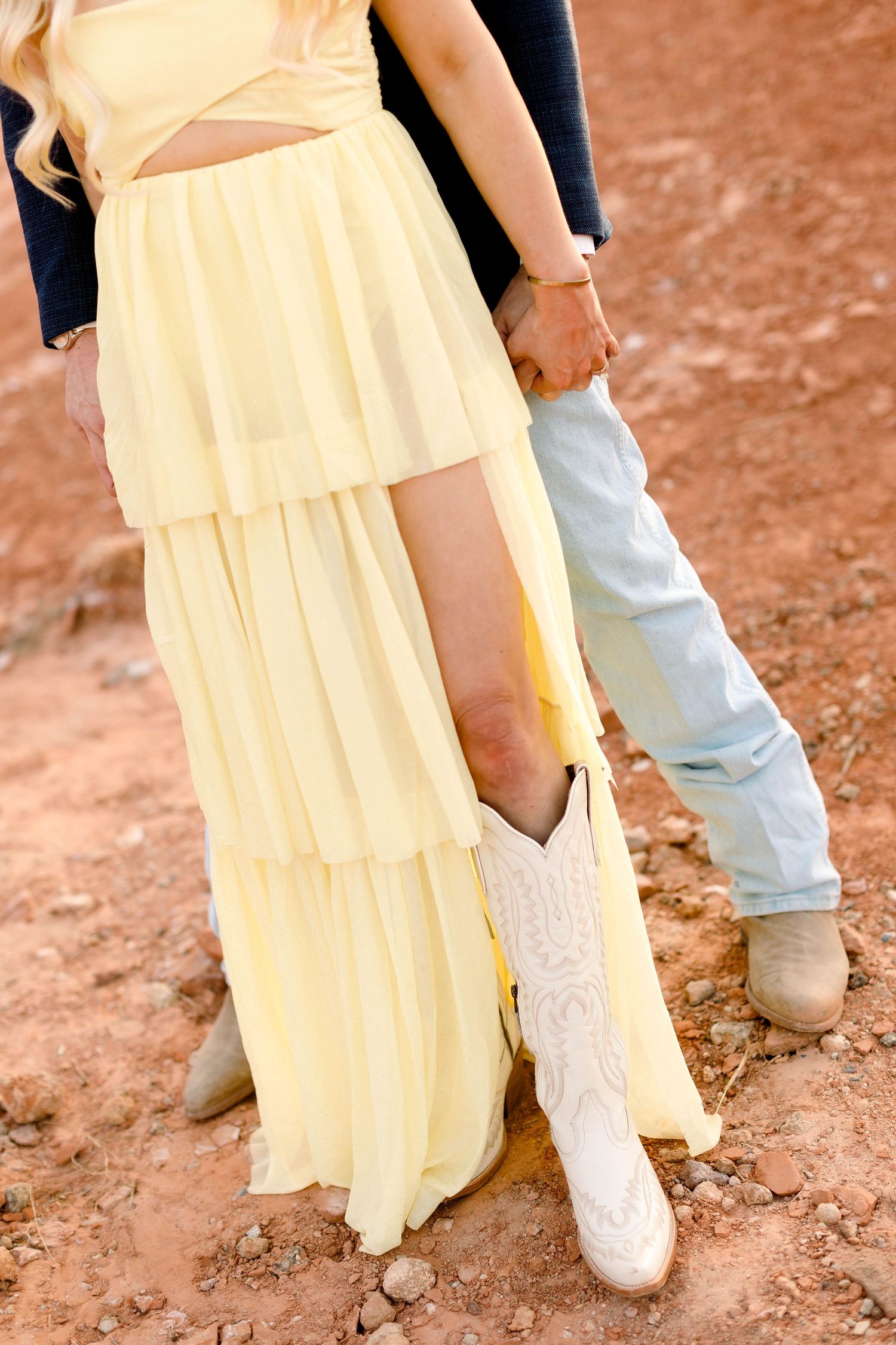 A close-up of white cowboy boots paired with a flowing yellow tiered dress against a red dirt background.