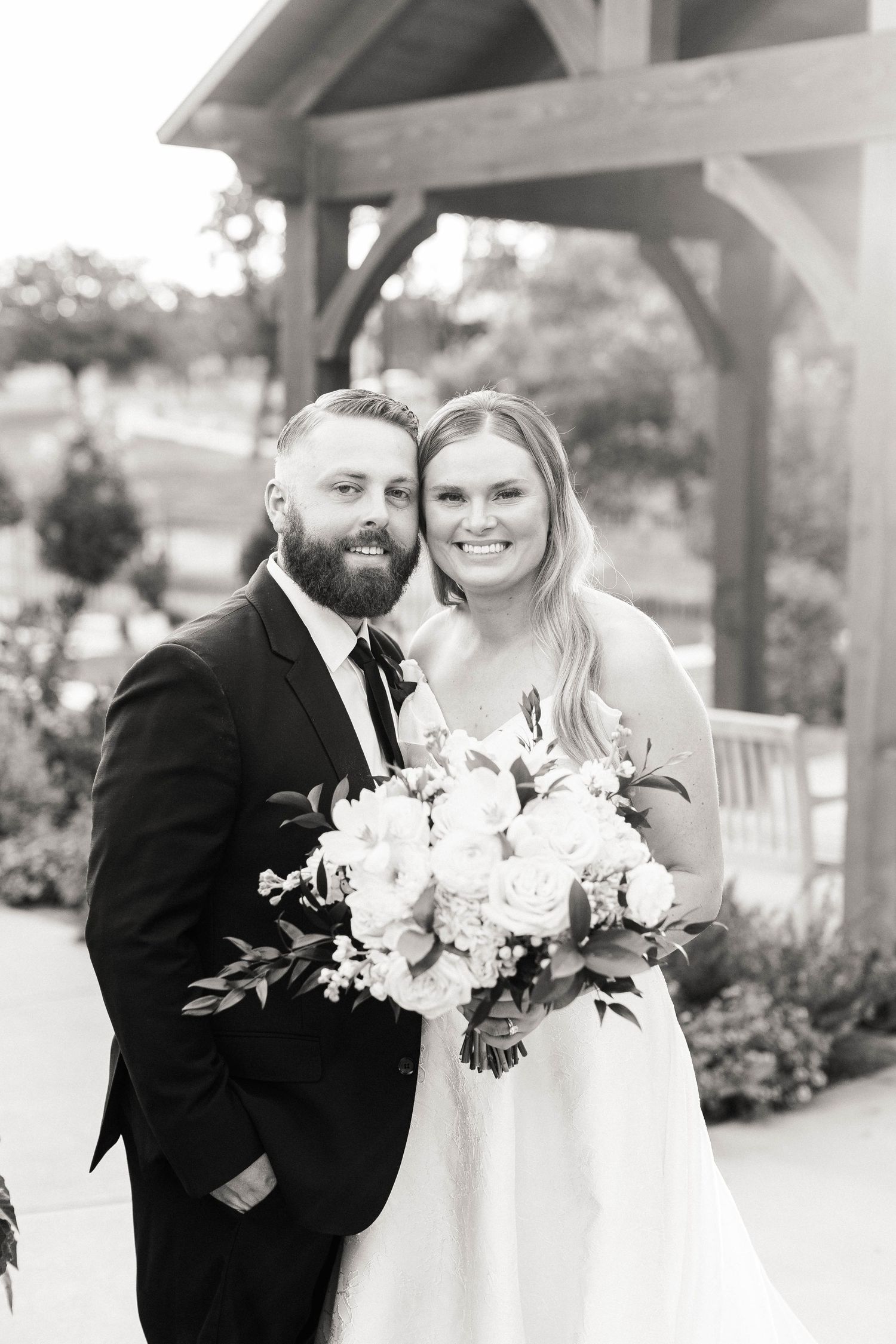 Black and white wedding portrait under wooden pergola with white floral bouquet.
