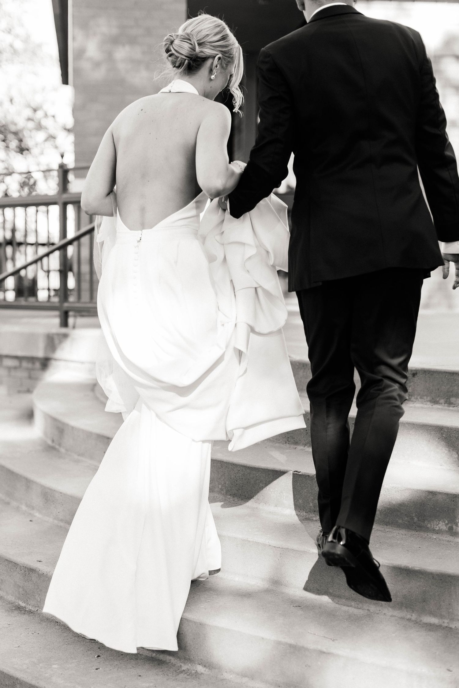 Black and white photo of couple ascending stone steps, showcasing the bride's backless dress.