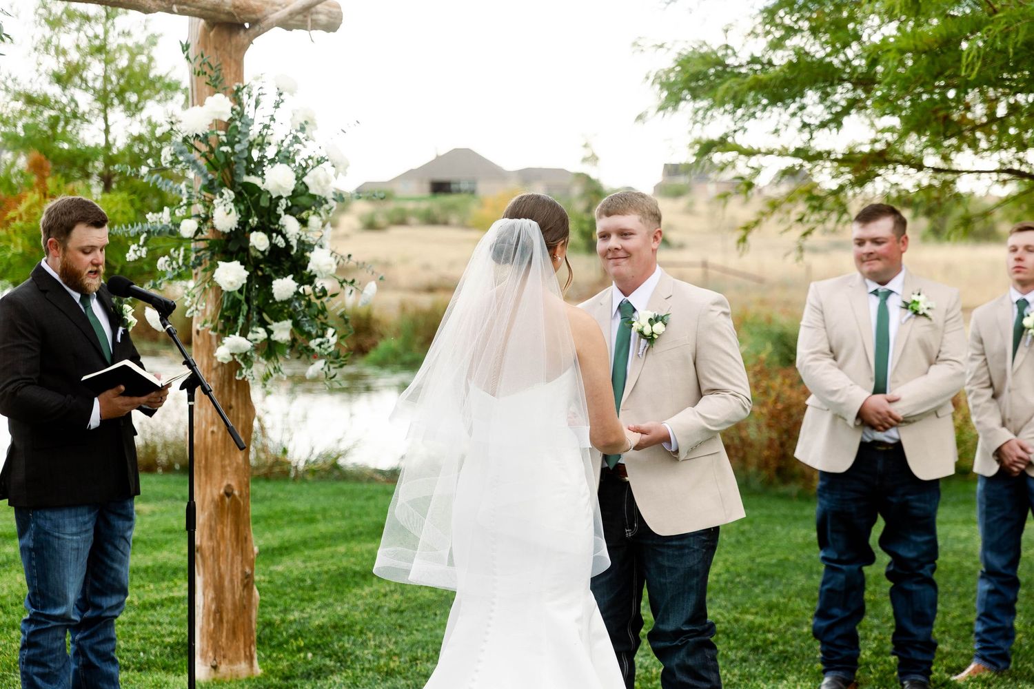 Wedding party poses under rustic wooden arch decorated with white floral arrangements.
