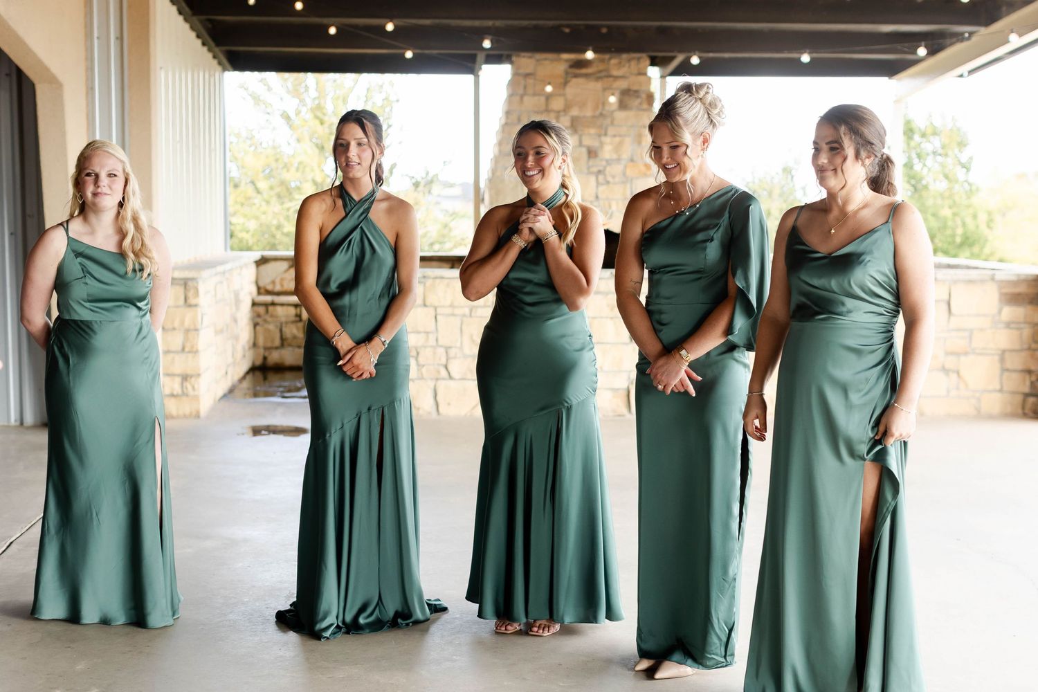 A row of bridesmaids in matching emerald green dresses pose together against a stone wall backdrop.