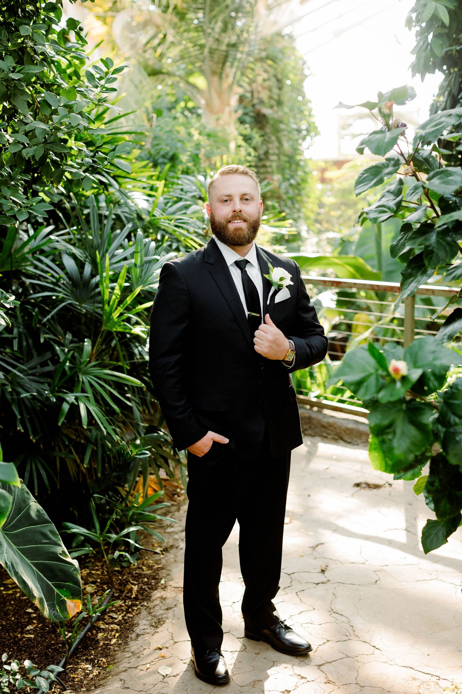 A person in a black suit poses playfully in a lush garden setting with tropical plants and stone pathways.