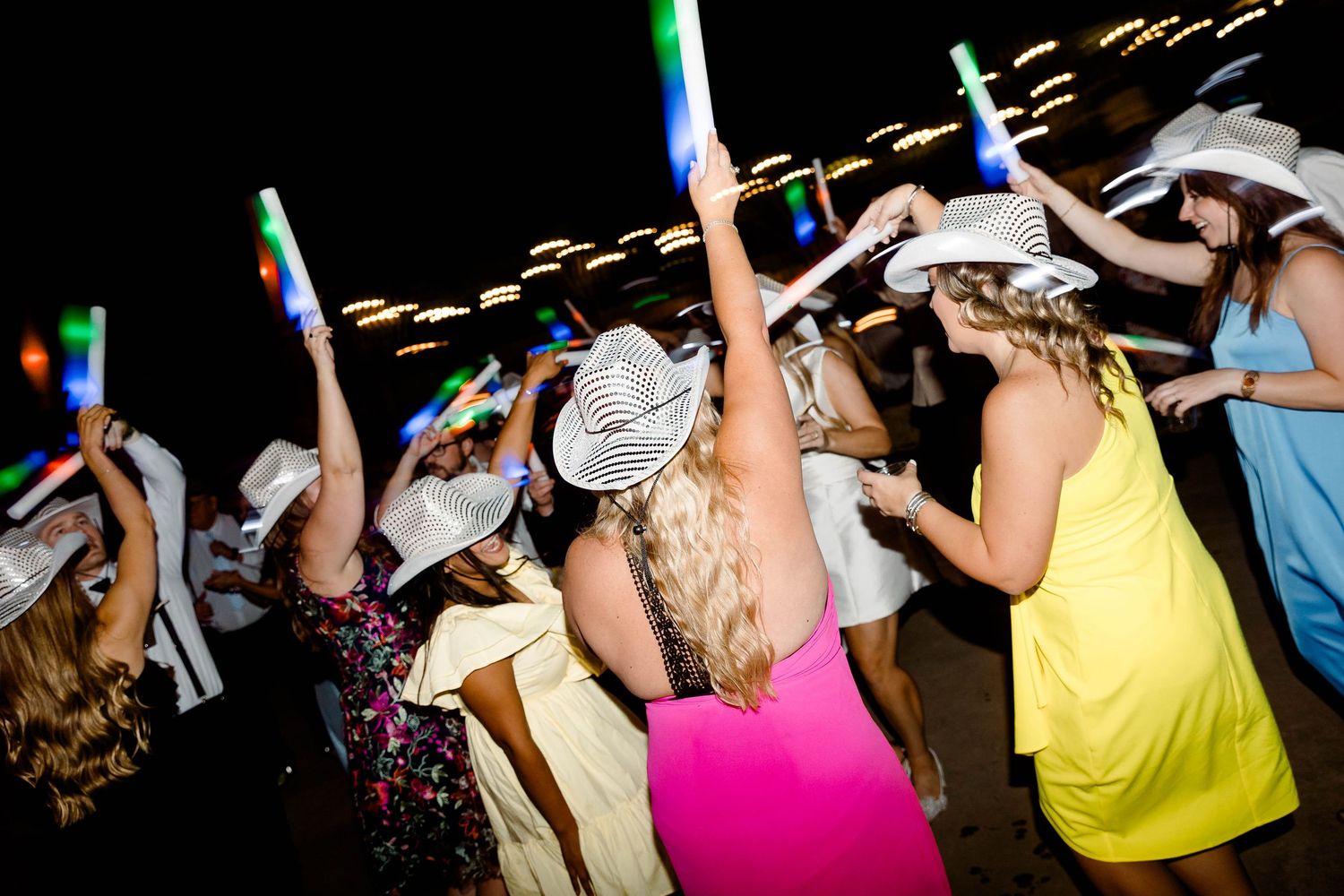 Party crowd dancing and celebrating with raised arms under colorful lights at a nighttime event.