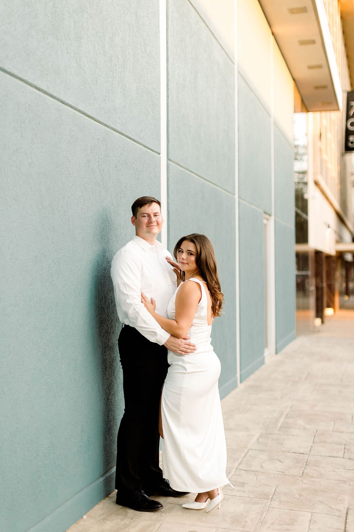 A romantic couple poses against a minimalist wall in a series of engagement photos.
