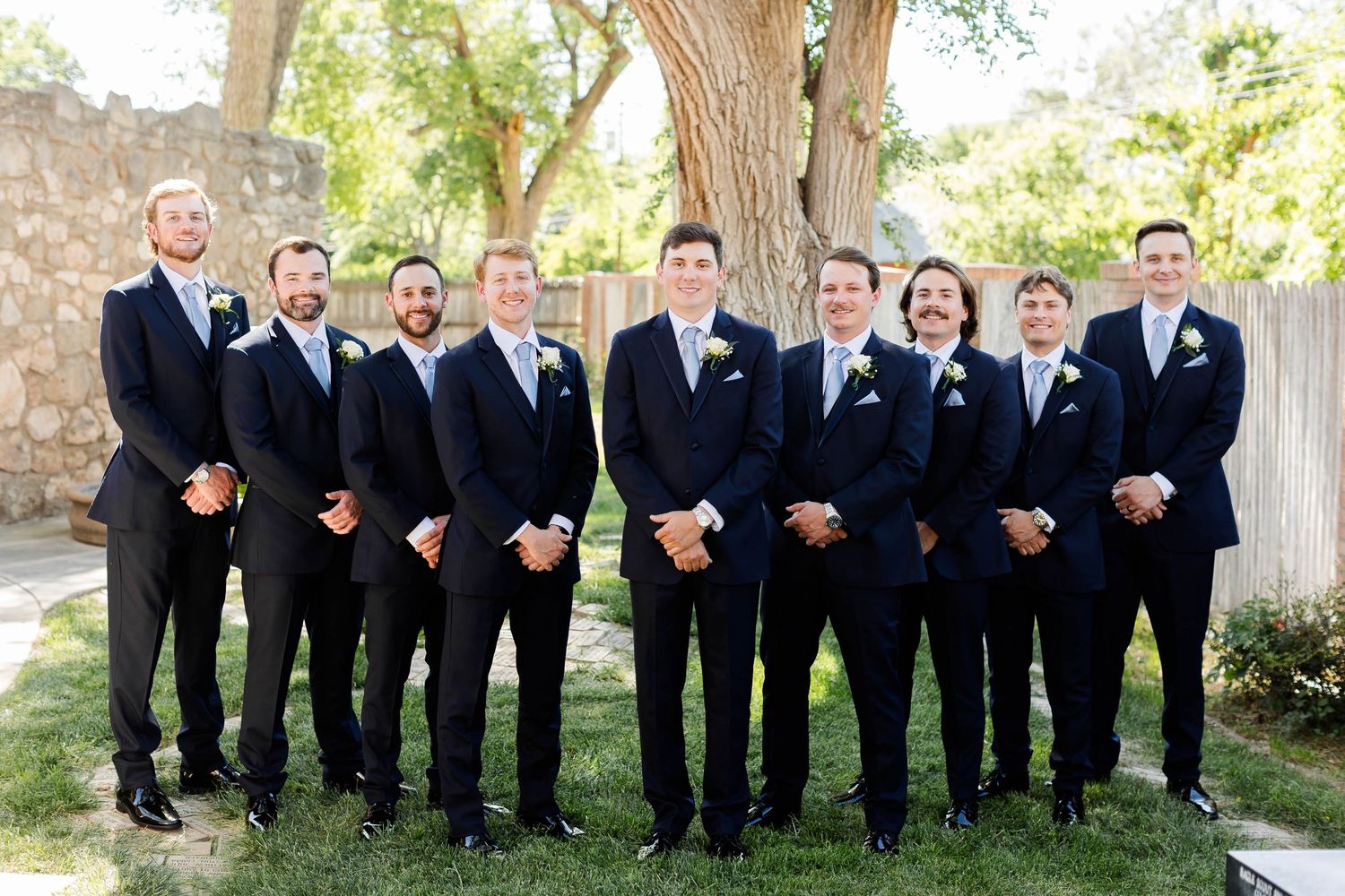 Groomsmen in matching black suits and ties pose together outdoors under a large tree.
