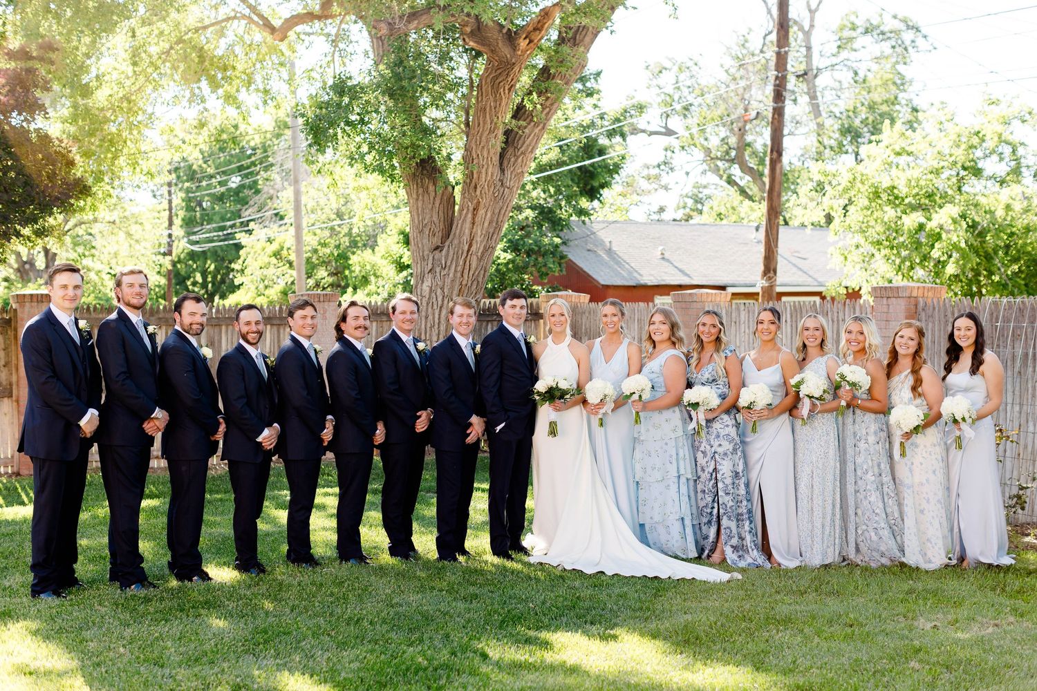 Large wedding party posed outdoors under trees in formal attire.