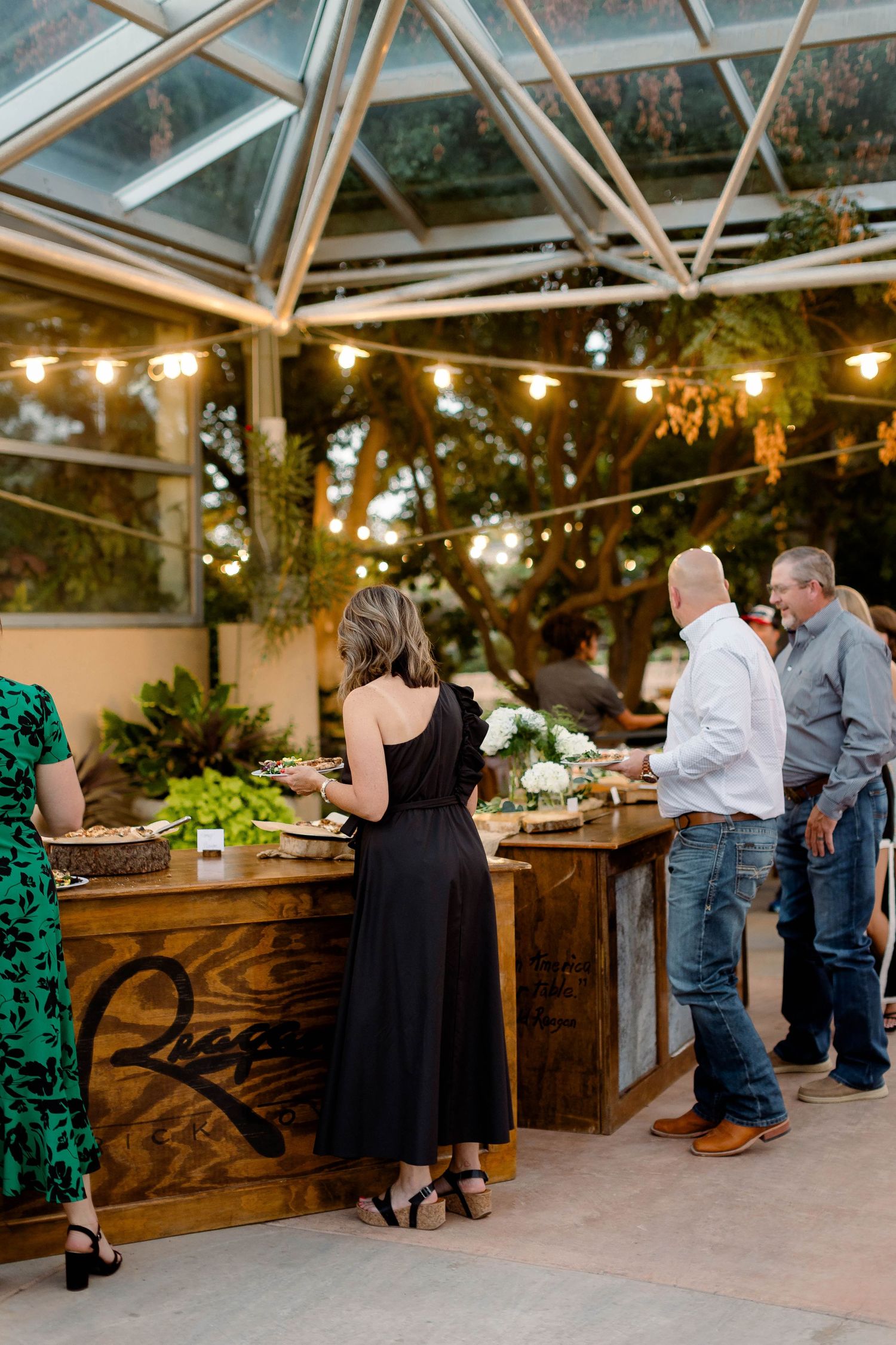 Guests mingling in a glass-roofed venue with wooden bar tables and ambient lighting.