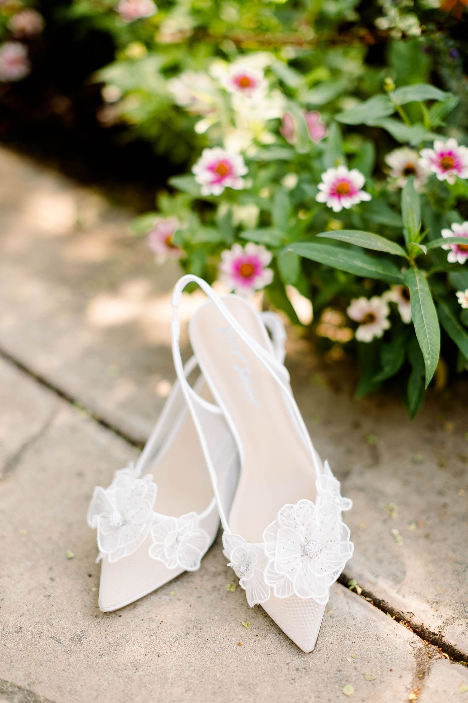 Elegant white lace bridal shoes displayed among pink garden flowers.