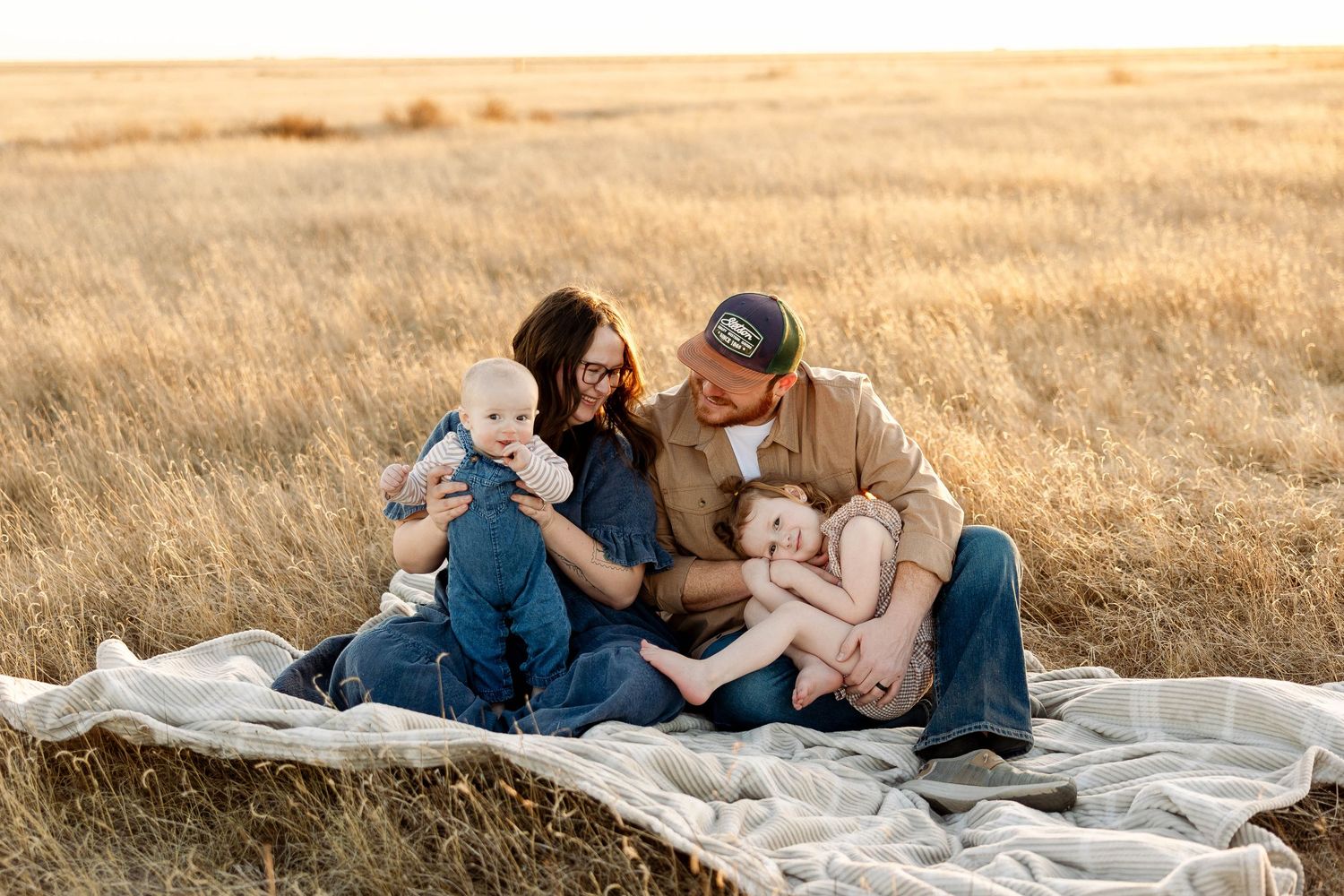 Family sitting together on blanket in golden wheat field during sunset.