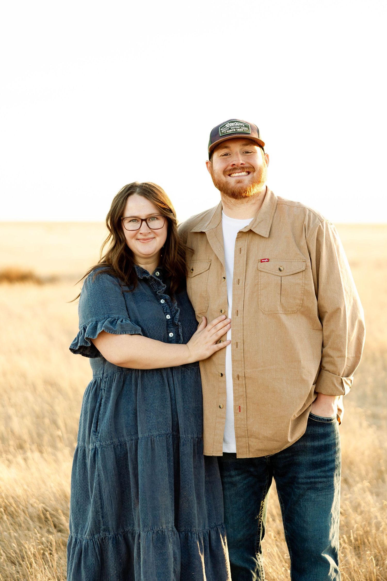 A couple stands together in a golden field during sunset wearing casual attire.