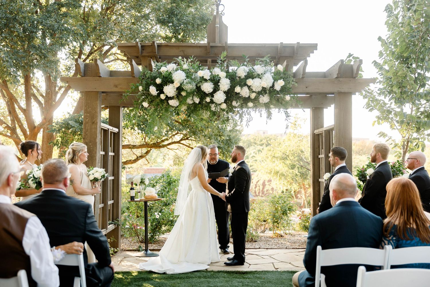 Wedding ceremony taking place in a garden setting with guests seated on white chairs.