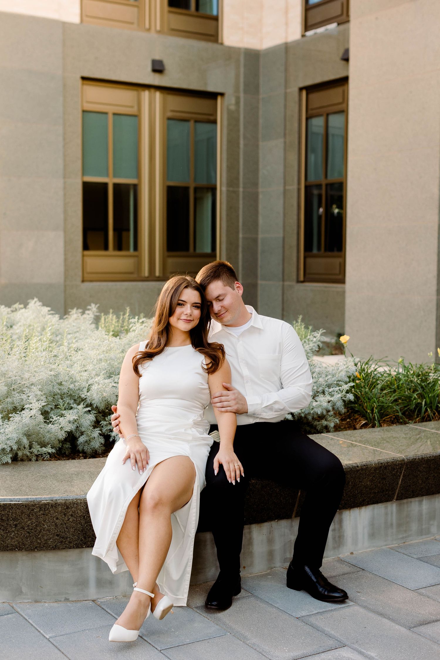 Couple in coordinating white outfits sits together on concrete steps near flowering bushes.