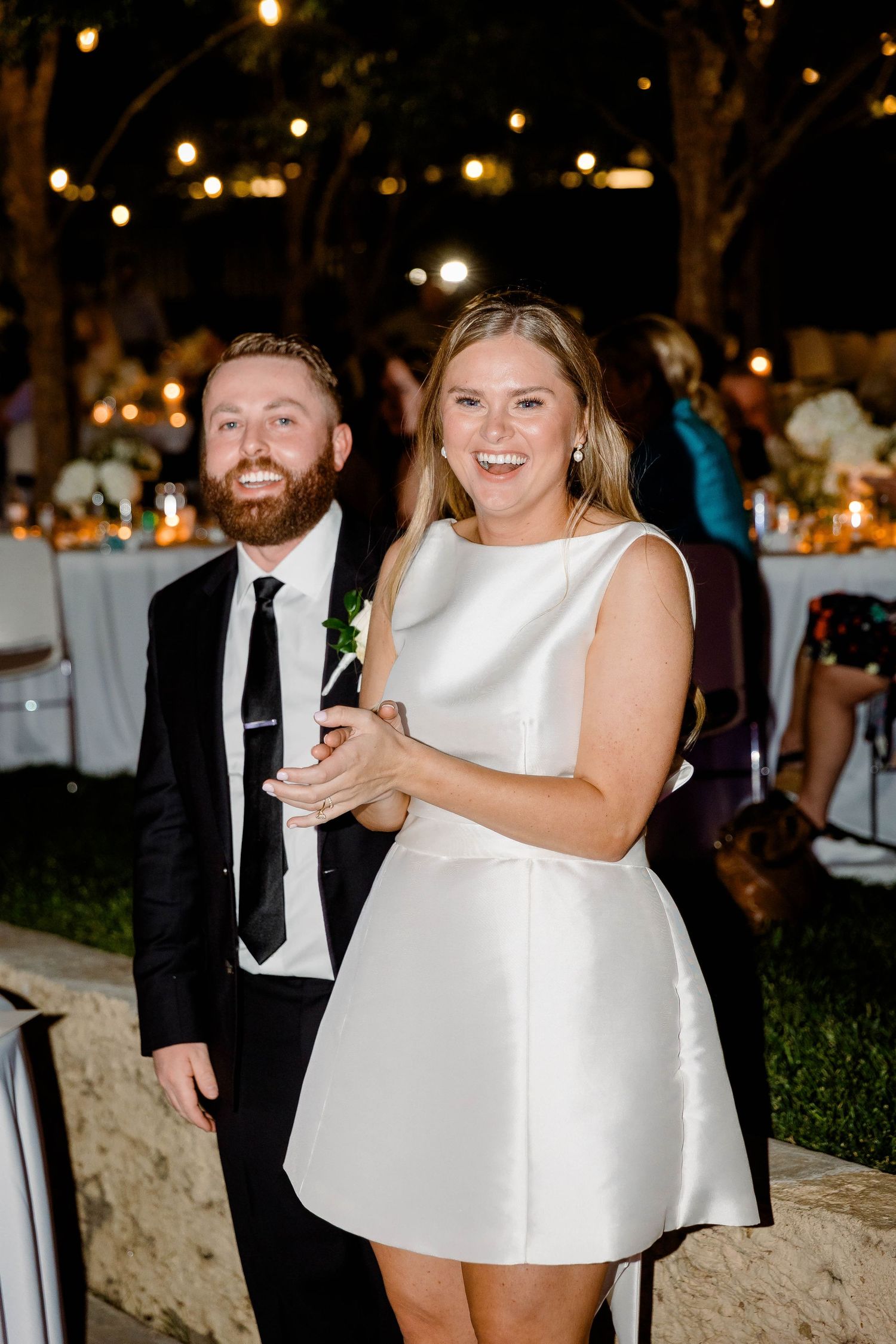 A bride and groom share a joyful moment during their wedding reception with twinkling lights in the background.