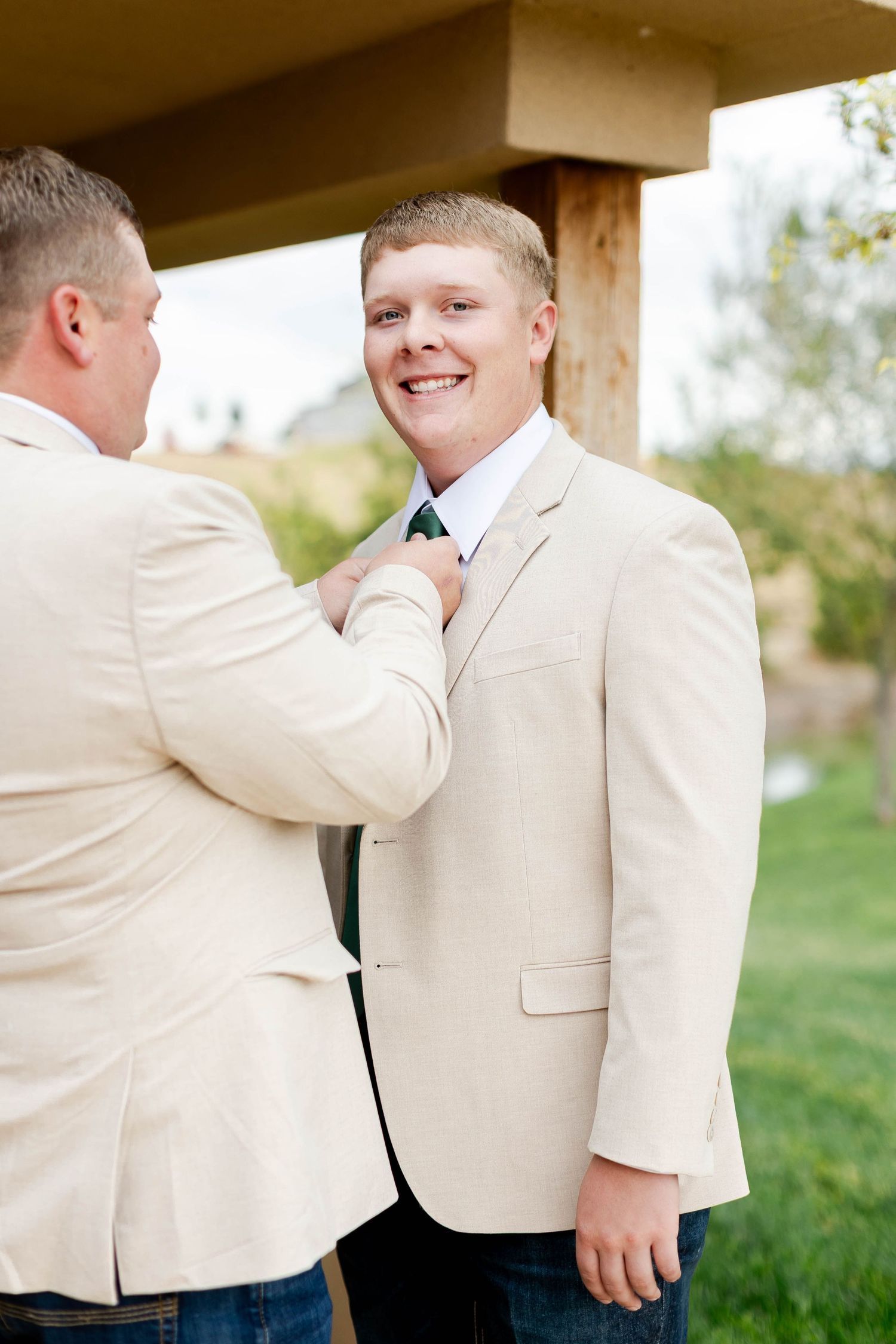 Someone helps adjust a tie on a person wearing a beige suit jacket at an outdoor event.