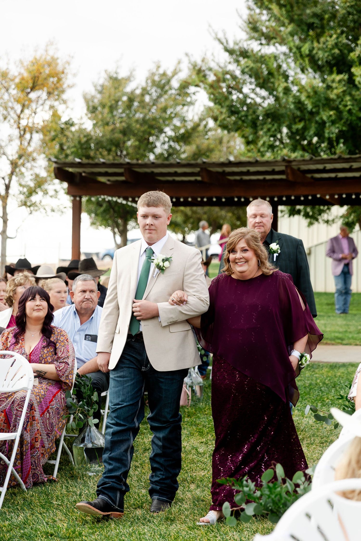 Wedding guests seated in white chairs on a grassy lawn with trees in the background.
