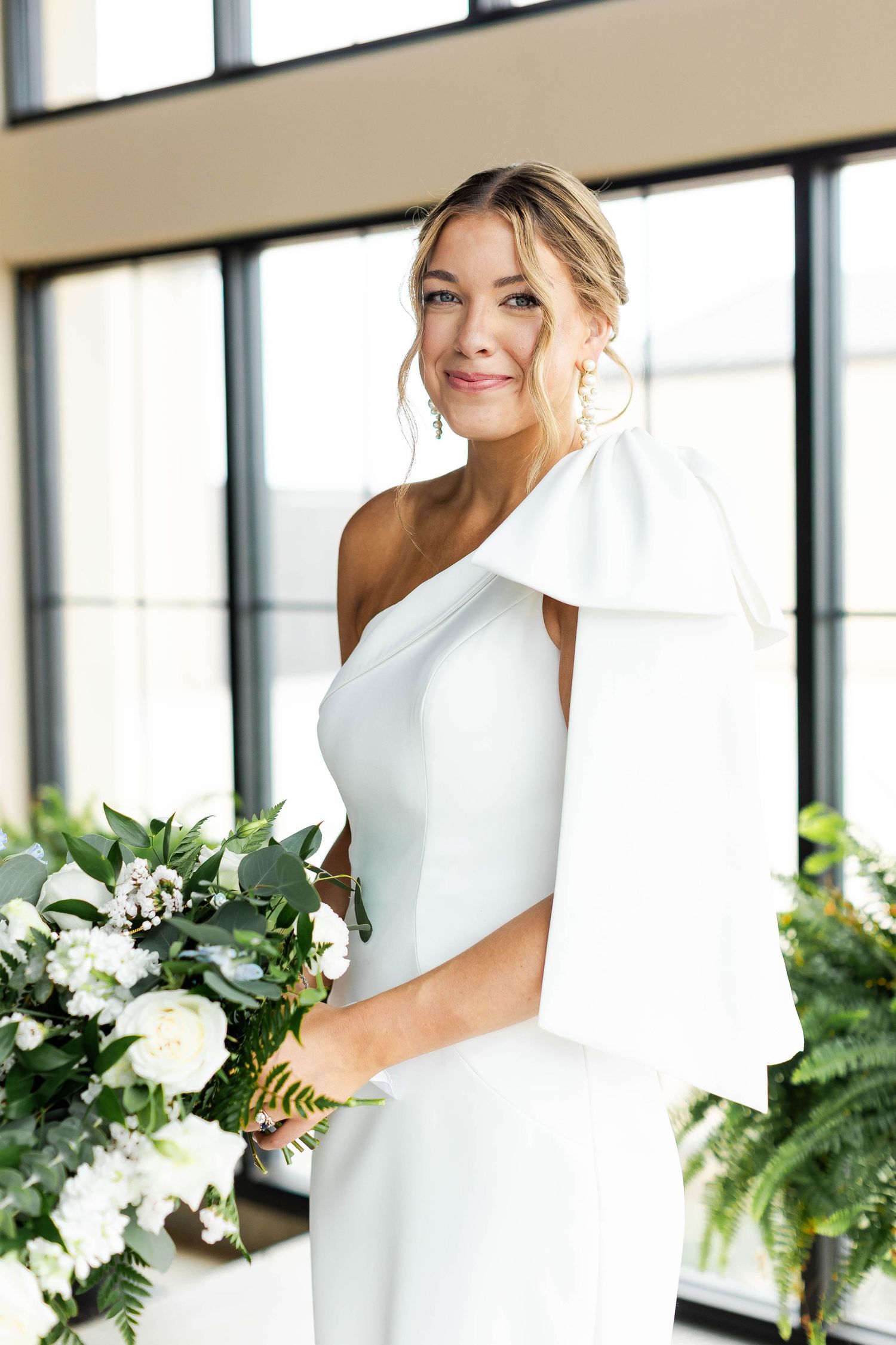 Modern one-shoulder wedding dress with dramatic bow detail and white floral bouquet.