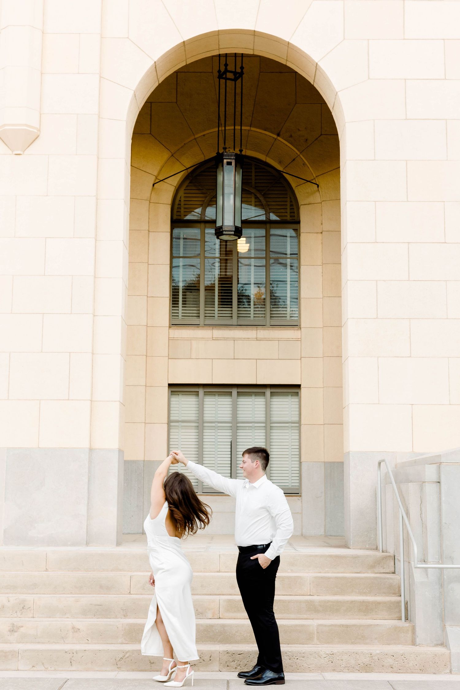 Couple dances and twirls on cream-colored stone steps with classic architecture.