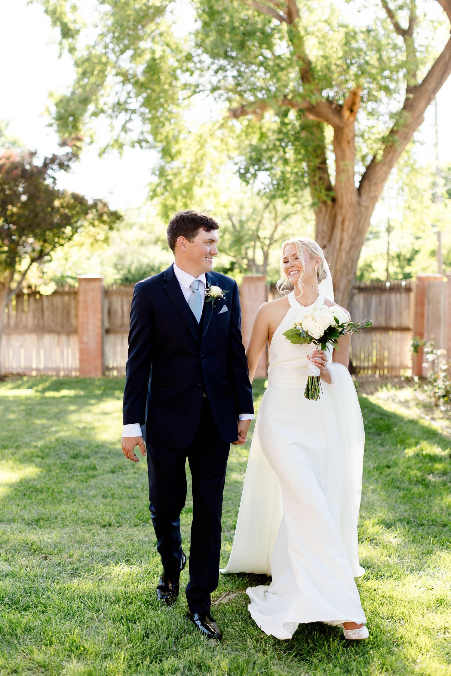 Happy newlyweds walking hand in hand on grass with beautiful summer trees behind them.