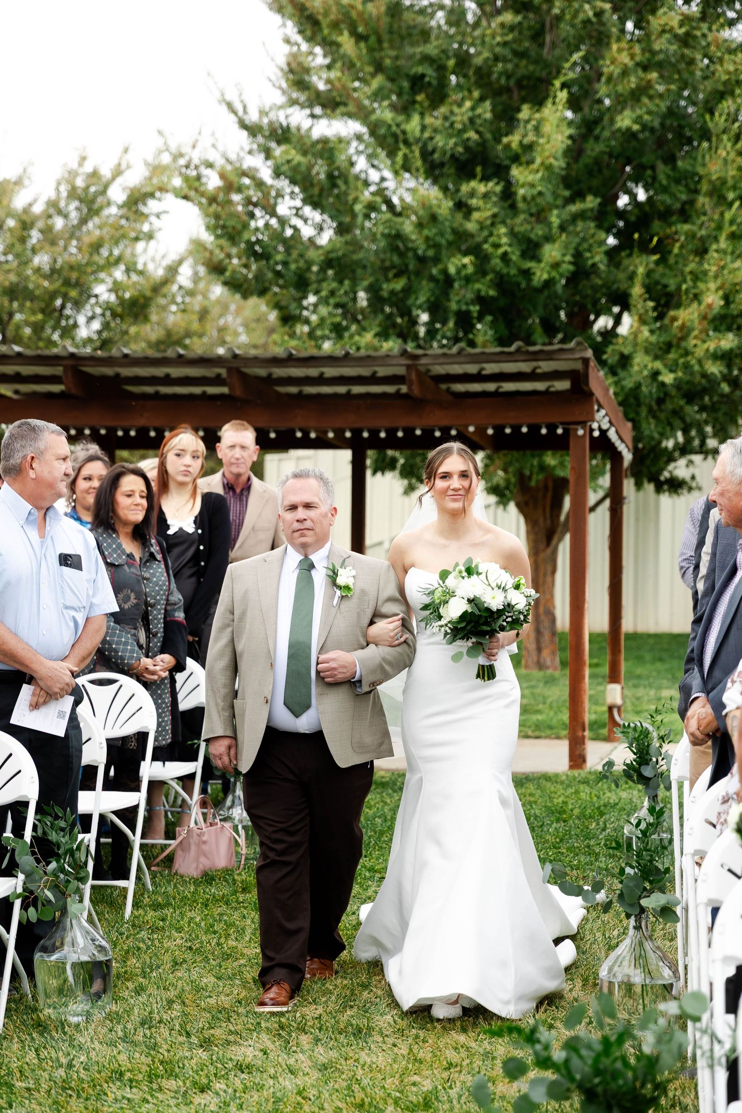An outdoor wedding ceremony processional takes place under a wooden pergola with string lights.