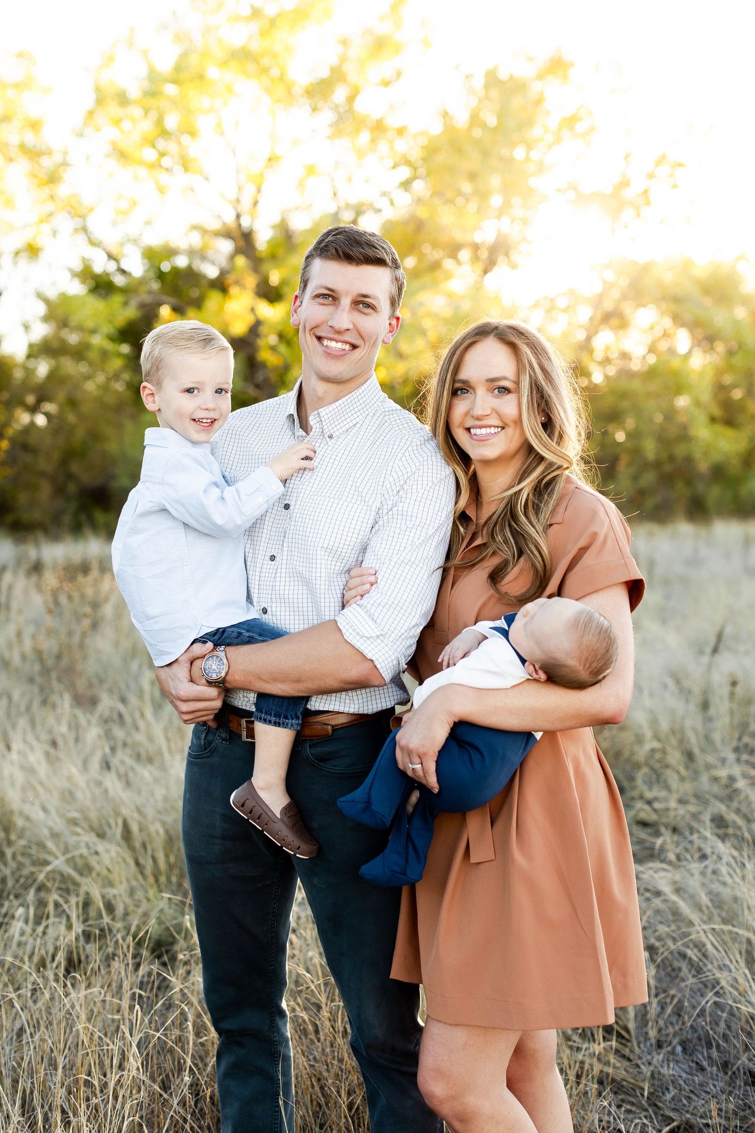 Family of three poses together in tall grass during golden sunset.