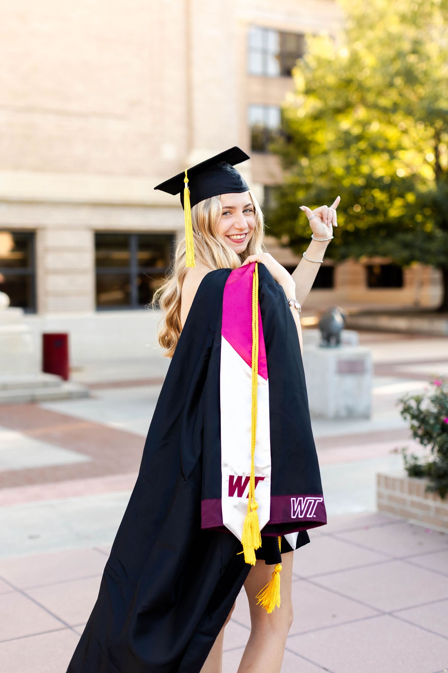 Graduate in academic regalia poses with diploma on campus steps.