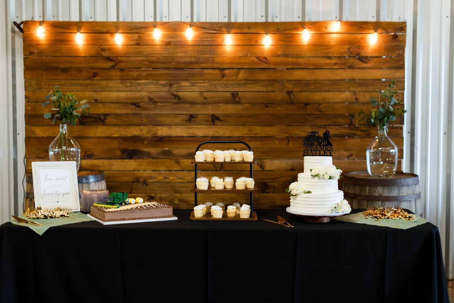 Wedding dessert table with white cake, cupcakes and treats displayed against rustic wood wall with string lights.