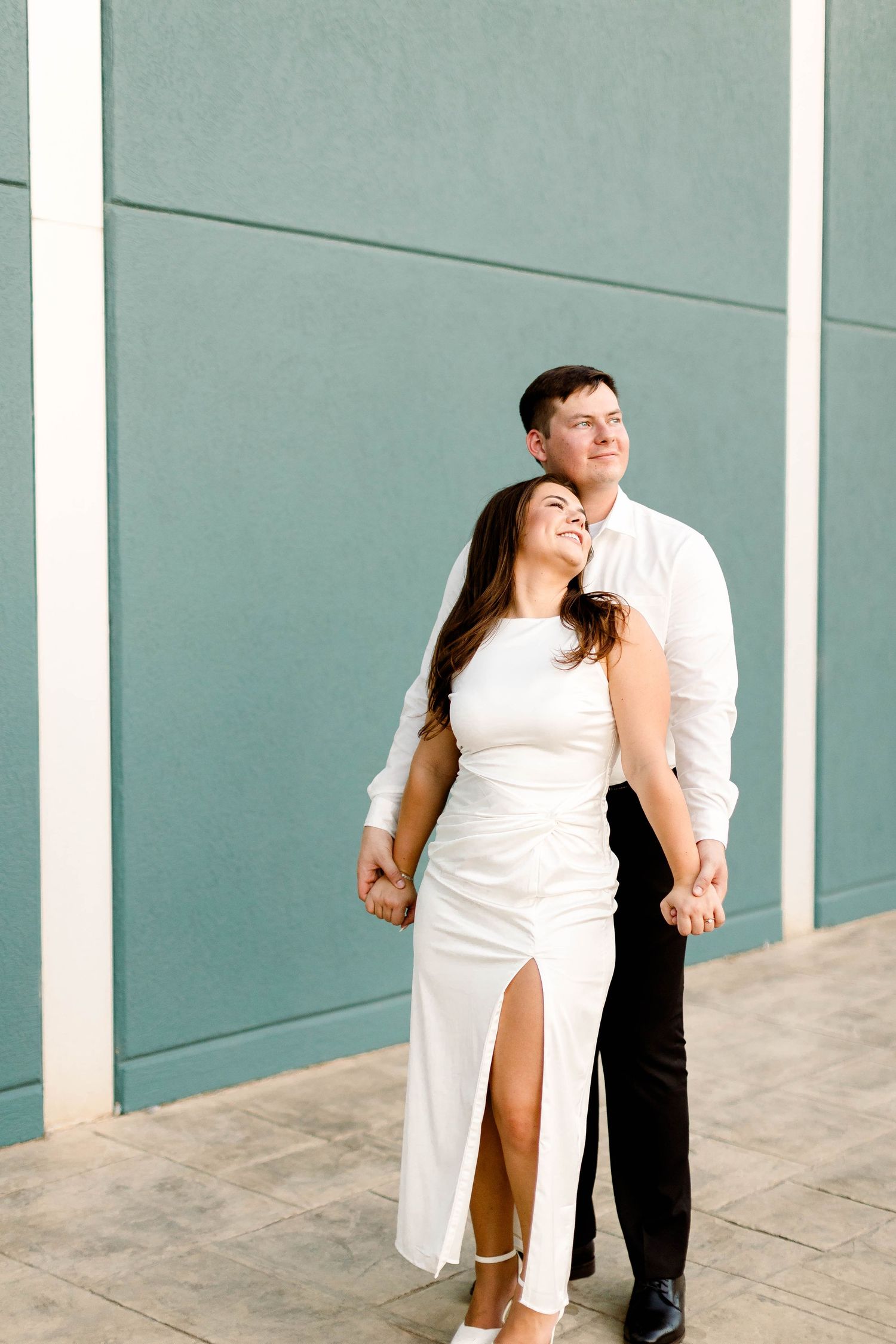 A couple shares candid laughing moments during their engagement photoshoot against a teal wall.