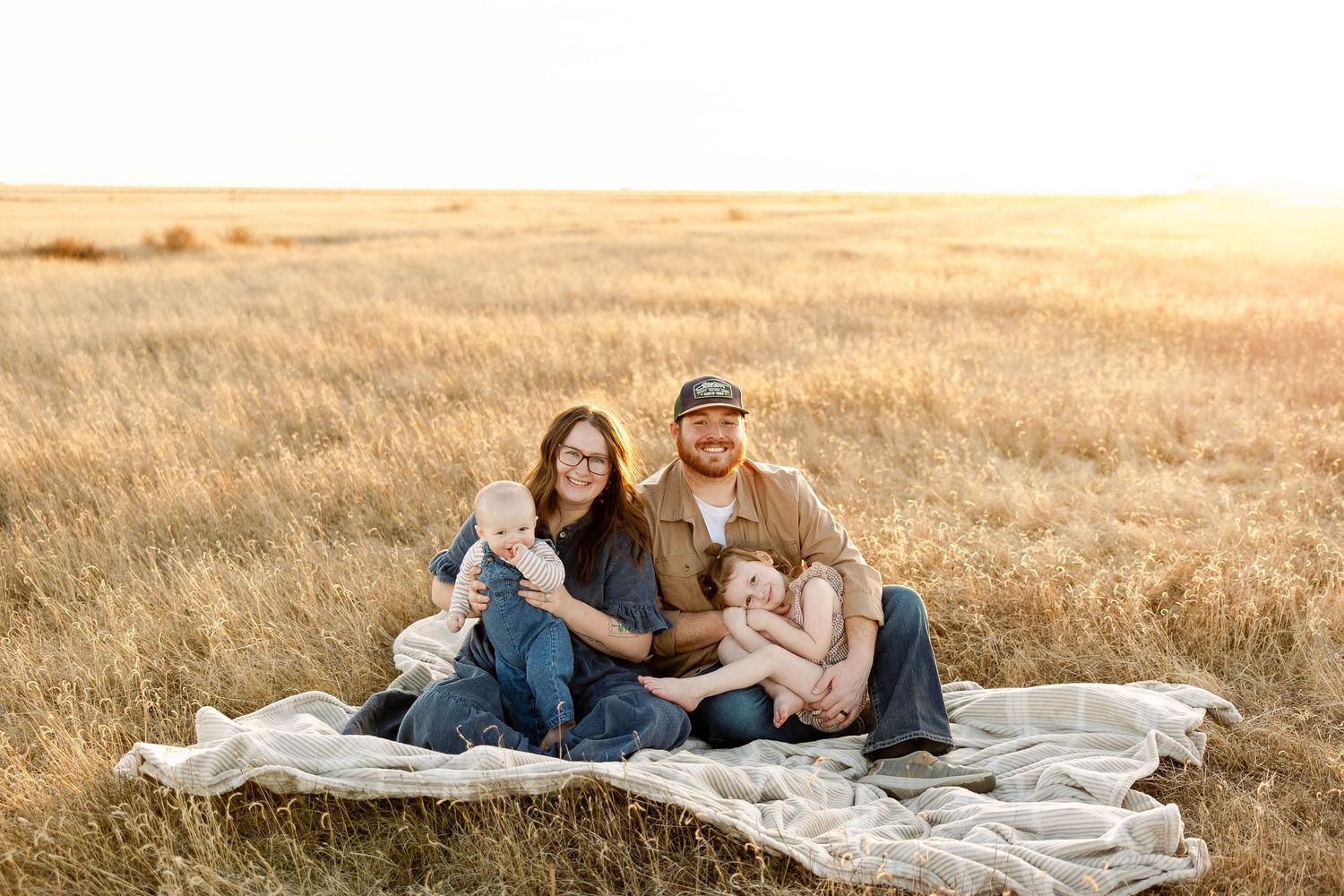 A couple sits on a white blanket in a golden wheat field during sunset.