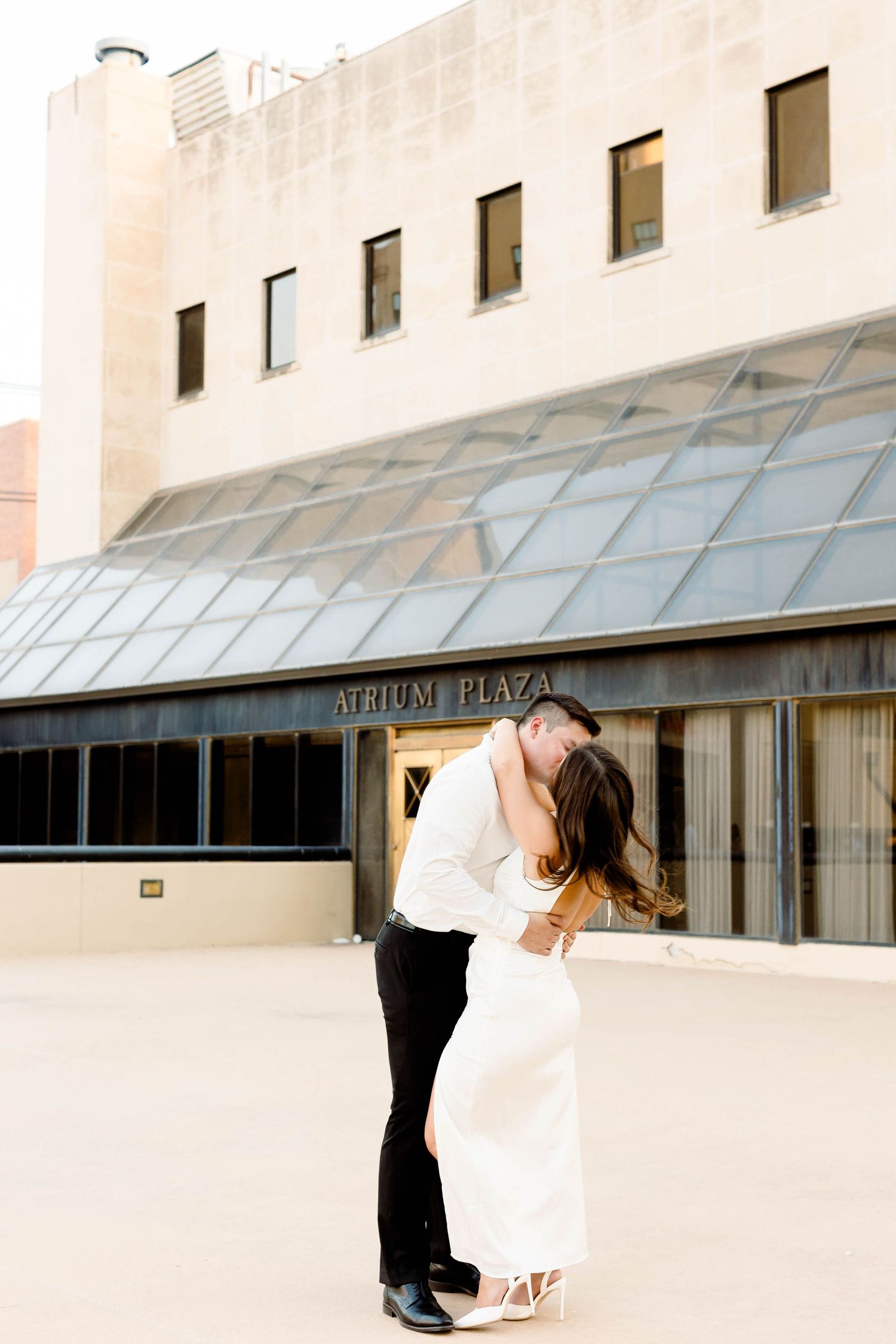 Romantic dance dip pose in front of modern glass building with Atrium Plaza signage.