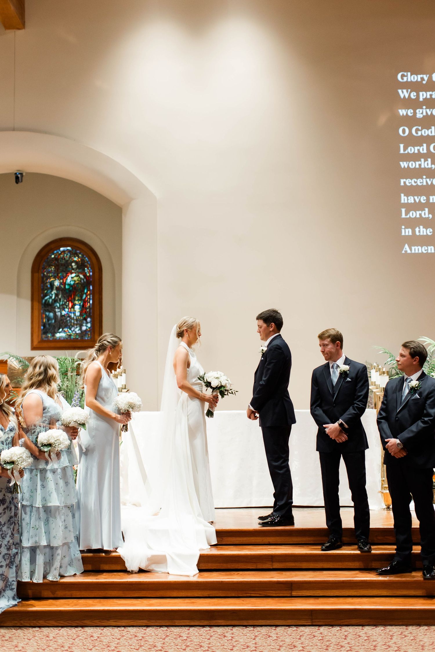 Wedding ceremony participants arranged on church altar with stained glass window in background.