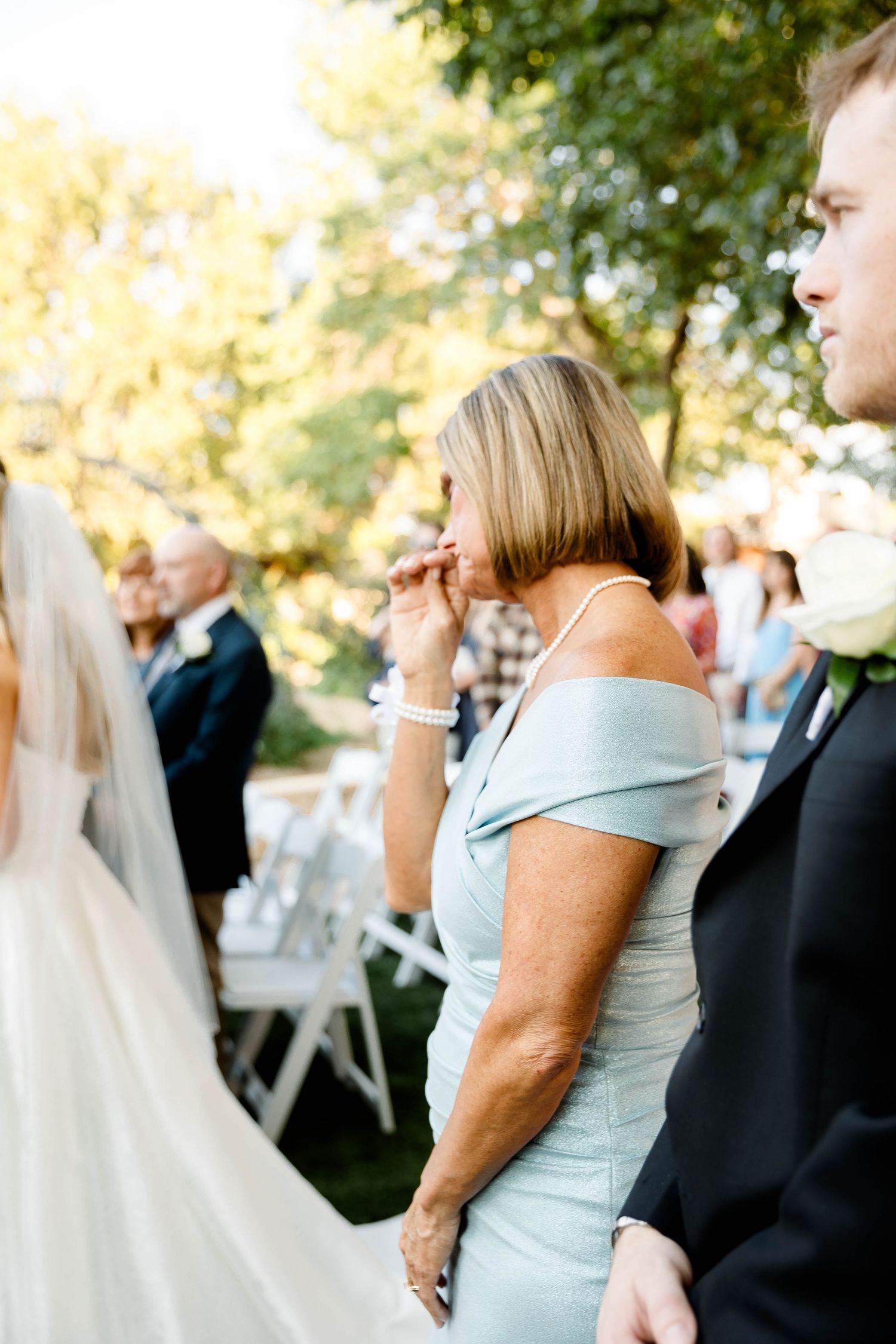 Wedding guests seated in white chairs during an outdoor ceremony under trees.