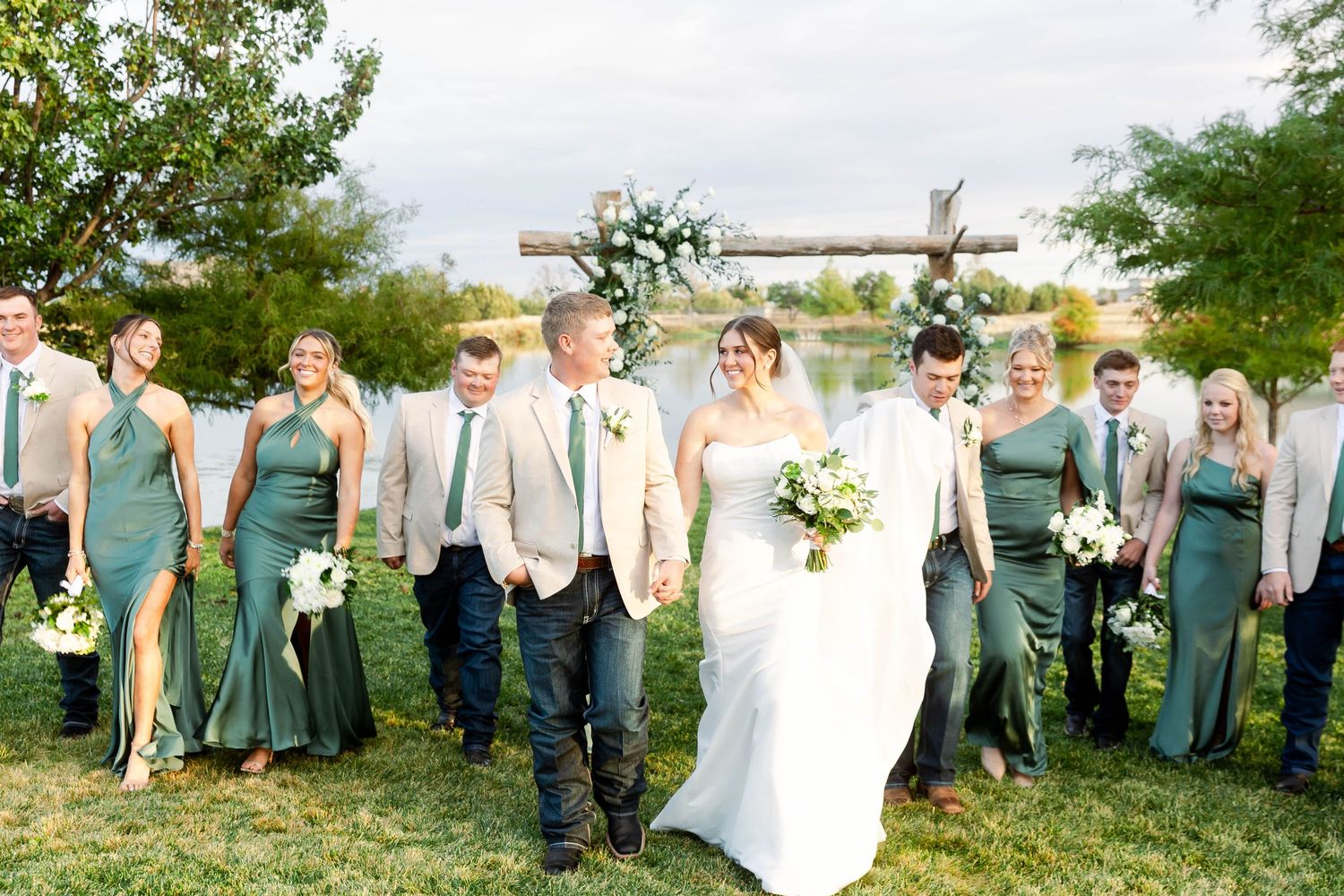 Wedding party in emerald green dresses and beige suits poses outdoors with a lake in the background.