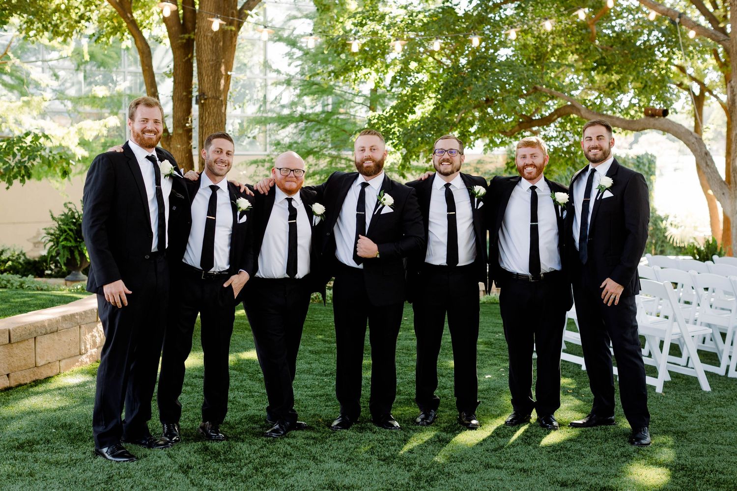 Wedding party members in black suits line up outdoors for a group photo.