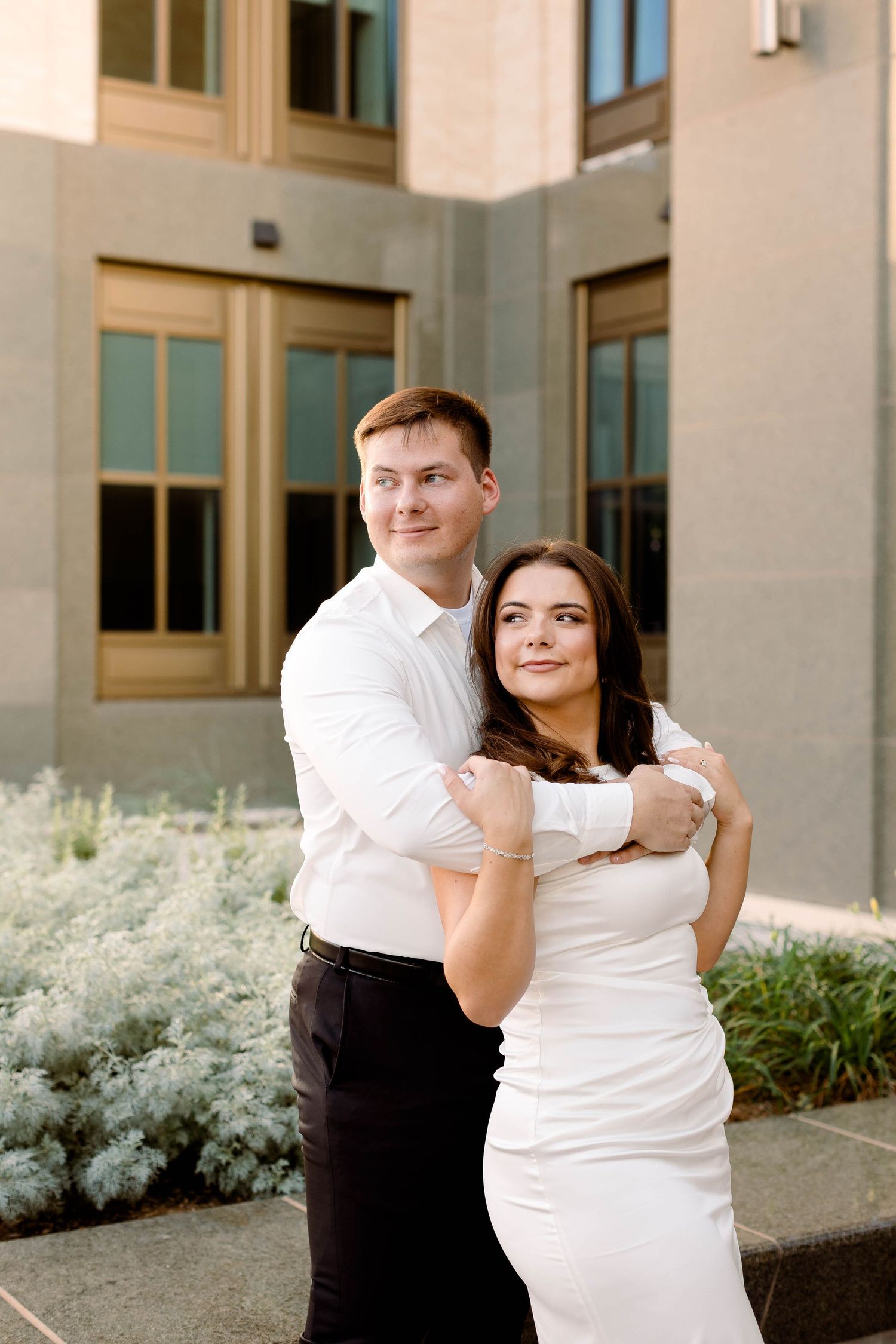 Elegant couple in white and black formal wear pose together in front of modern building.