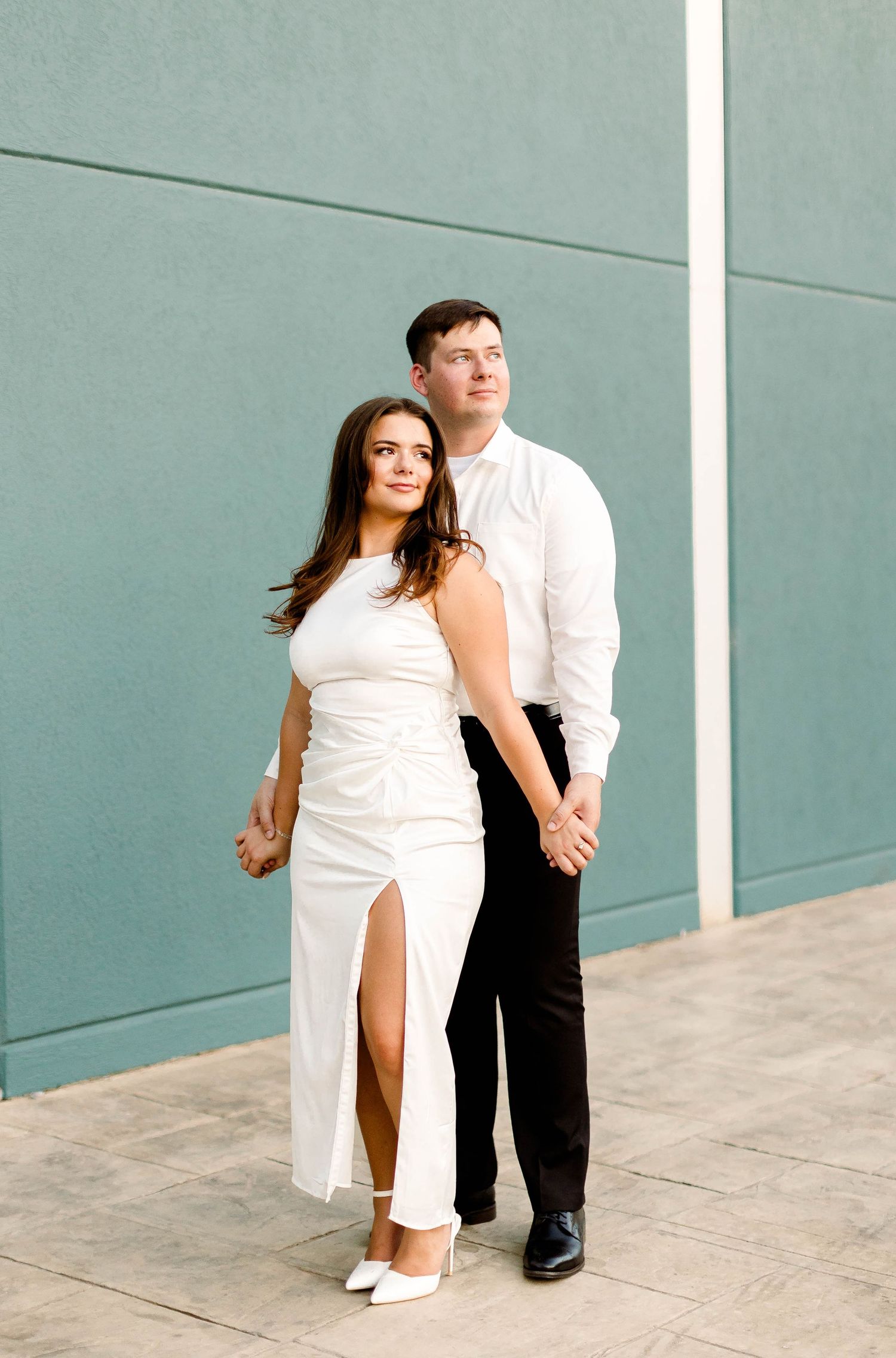 An engaged couple in formal attire poses playfully against a blue-gray wall.