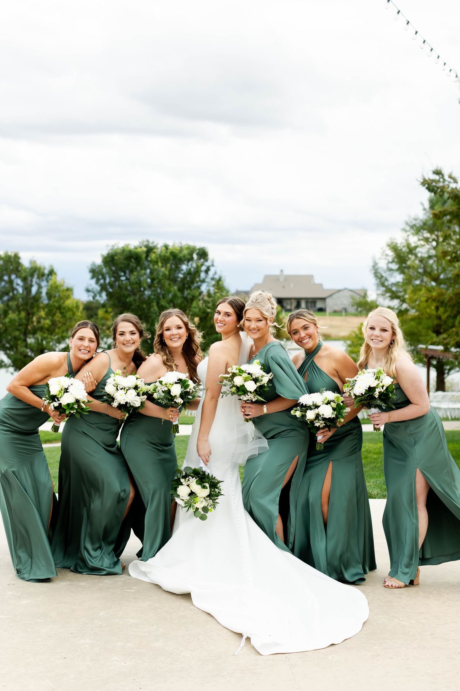 Bridal party in emerald green dresses poses playfully in a line on outdoor steps with white bouquets.