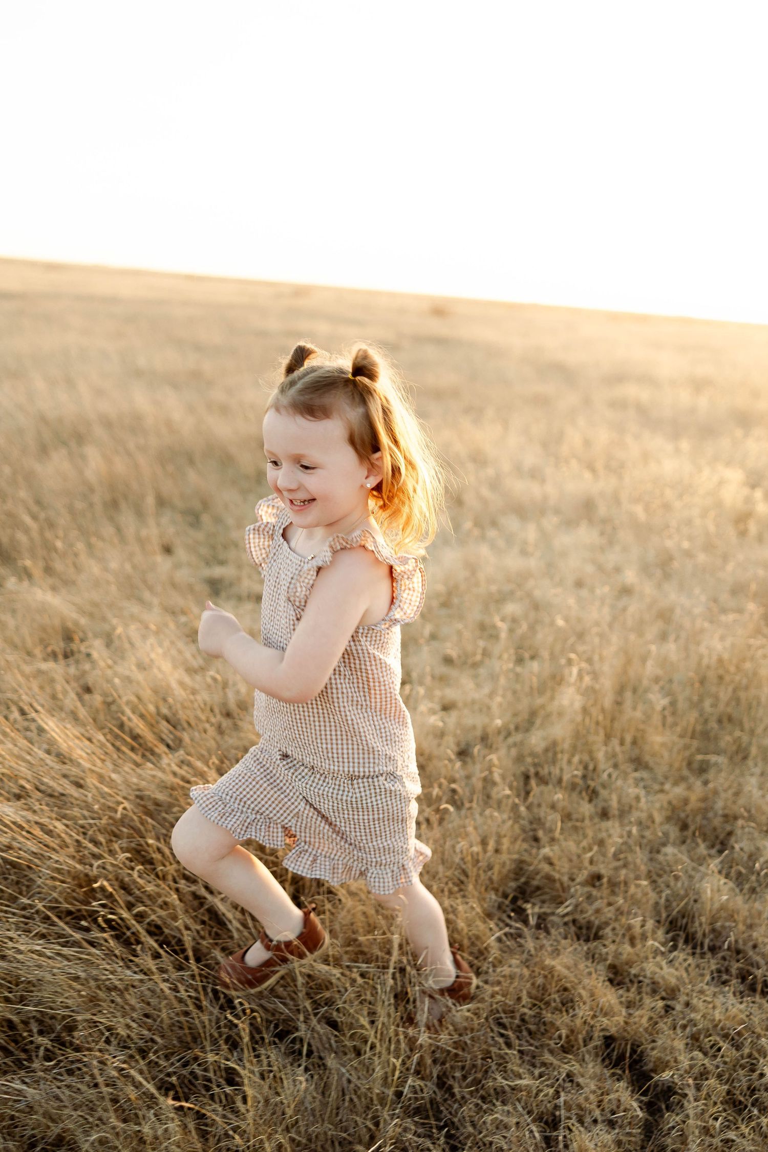 A young child in a gingham dress plays joyfully in a sunlit golden field during sunset.