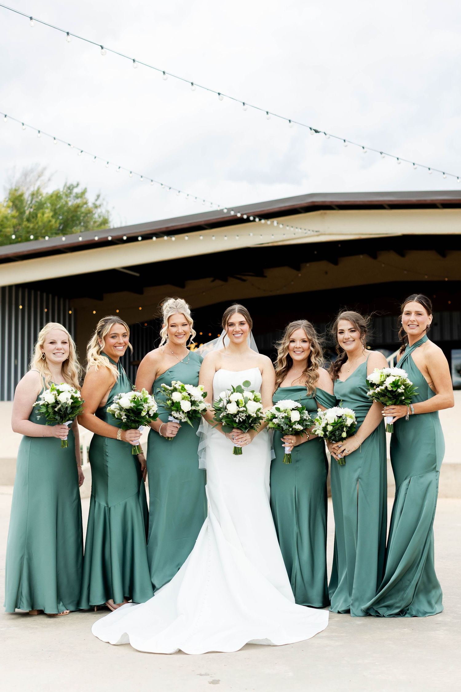 Wedding party in matching sage green dresses and white bouquets pose together against modern building exterior.