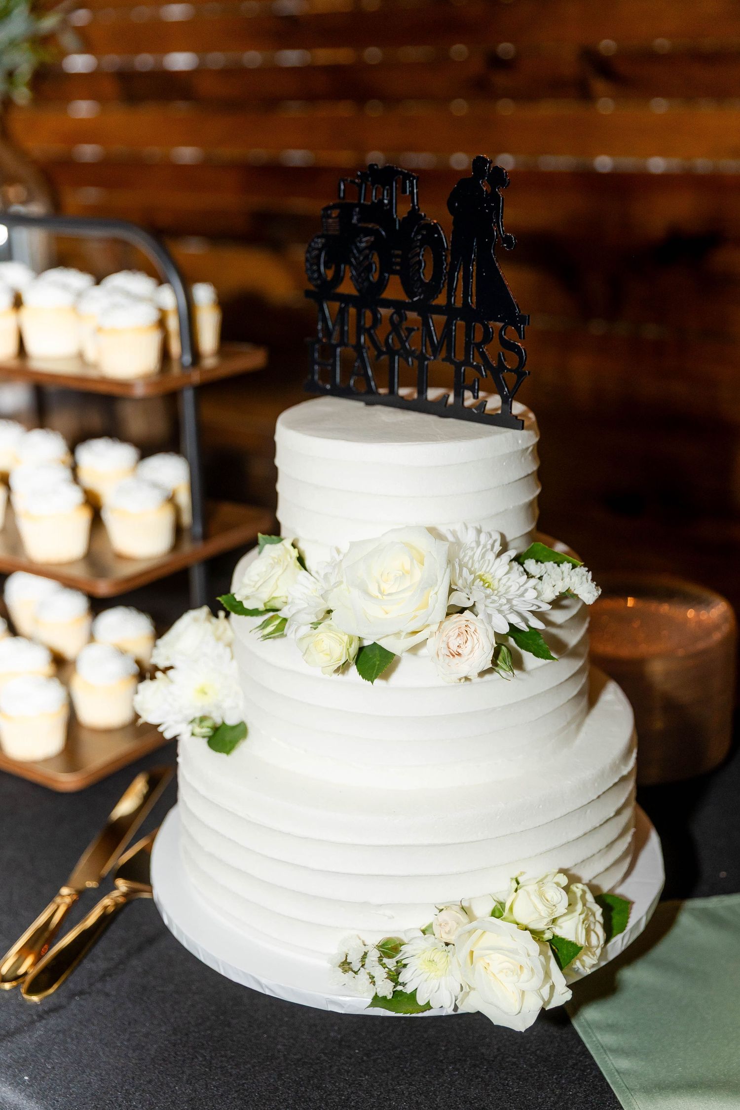White wedding cake with delicate floral decorations and a silhouette cake topper displayed in a rustic venue.